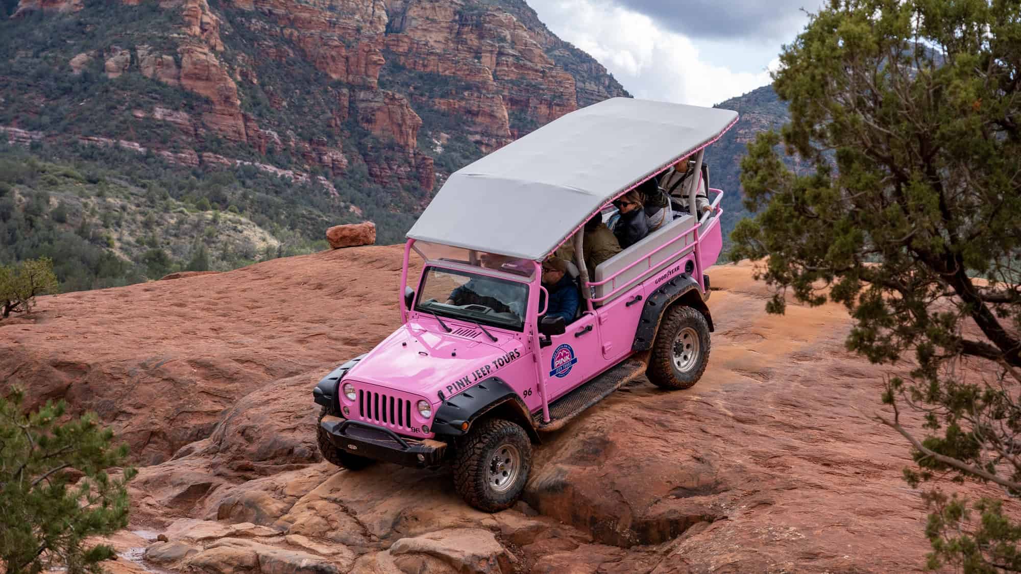 A bright pink off-road tour jeep with passengers navigates rugged red rock terrain in Sedona, surrounded by dramatic cliffs and scattered greenery.