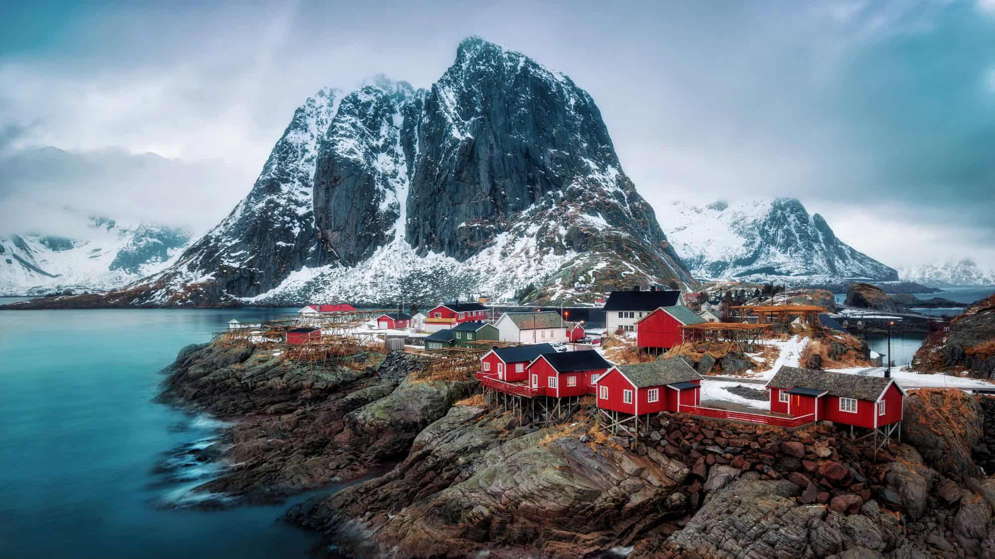 Red fishing cabins perched on rocky shores contrast against the dramatic snowy mountains of Reine, a picturesque village in Norway's Lofoten Islands under a moody winter sky.