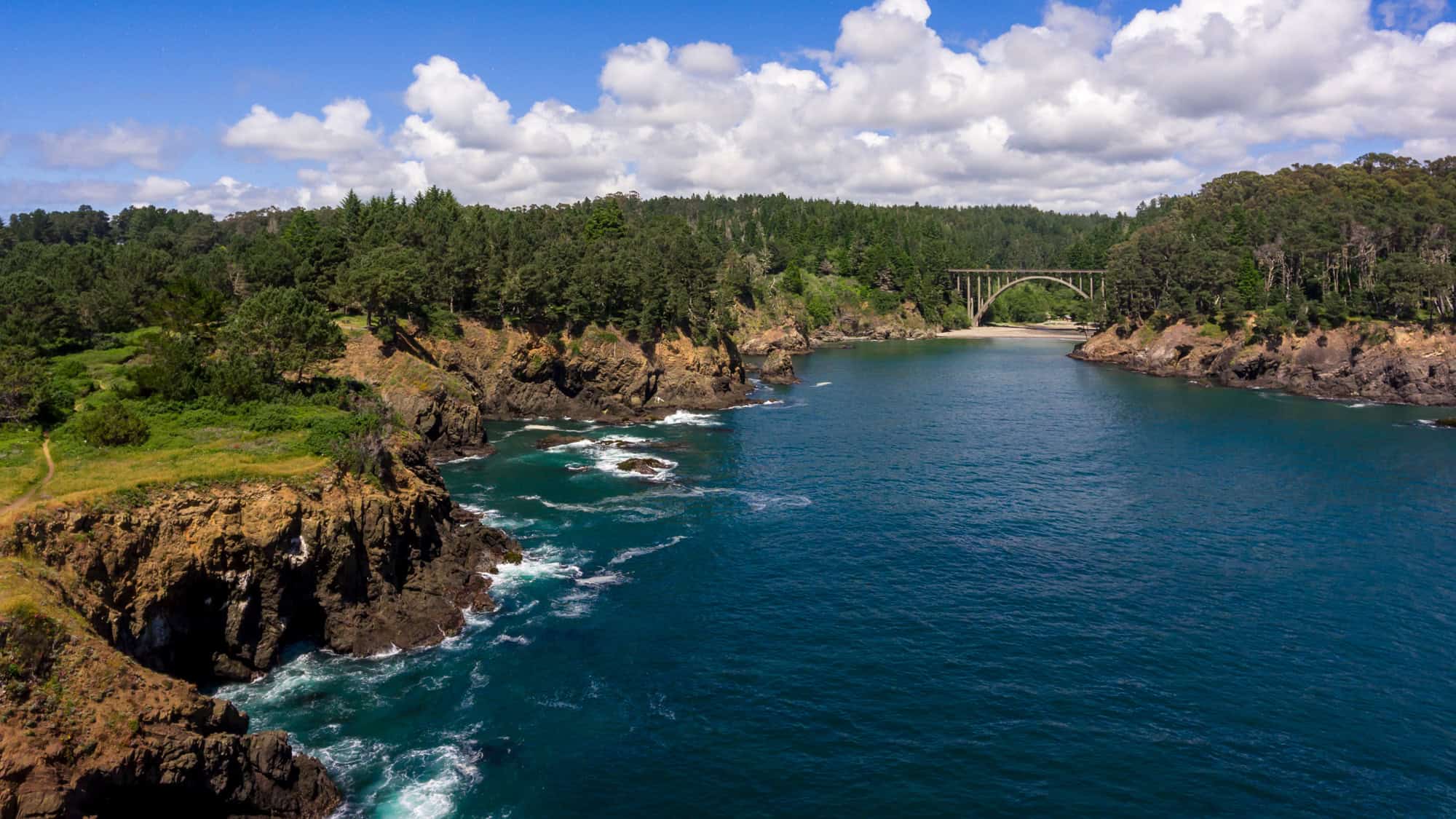 A rugged coastal inlet bordered by pine-covered cliffs, with a scenic arched bridge in the background and turquoise waves crashing below under a bright, partly cloudy sky.
