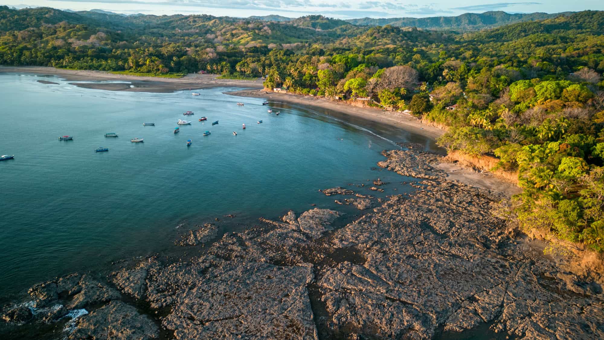 An aerial view of a quiet, crescent-shaped bay in Panama with fishing boats anchored offshore, a tree-lined shoreline, and lush green forest stretching into the hills.