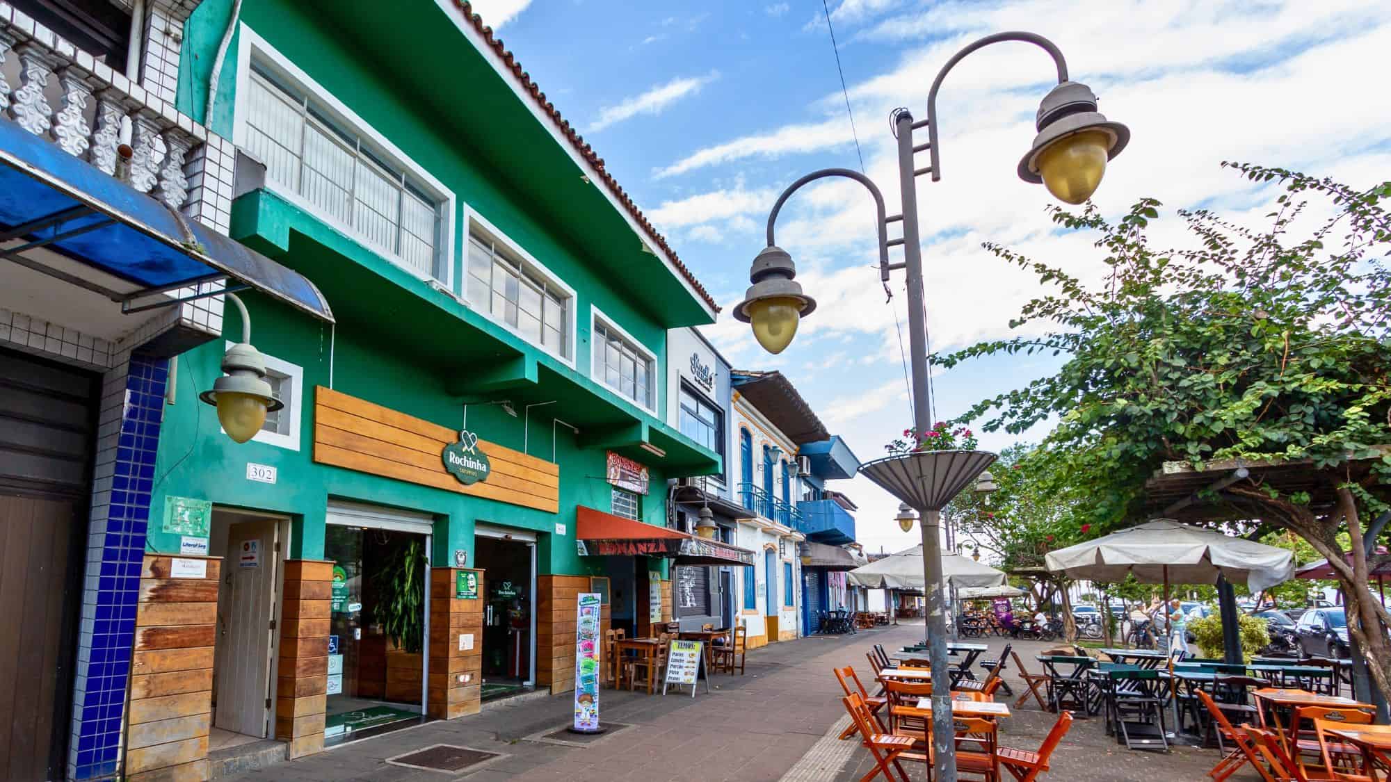 A vibrant street in São Sebastião, Brazil features colorful buildings, outdoor seating with wooden tables and umbrellas, and tropical greenery. The area exudes a lively, laid-back atmosphere with a coastal charm.