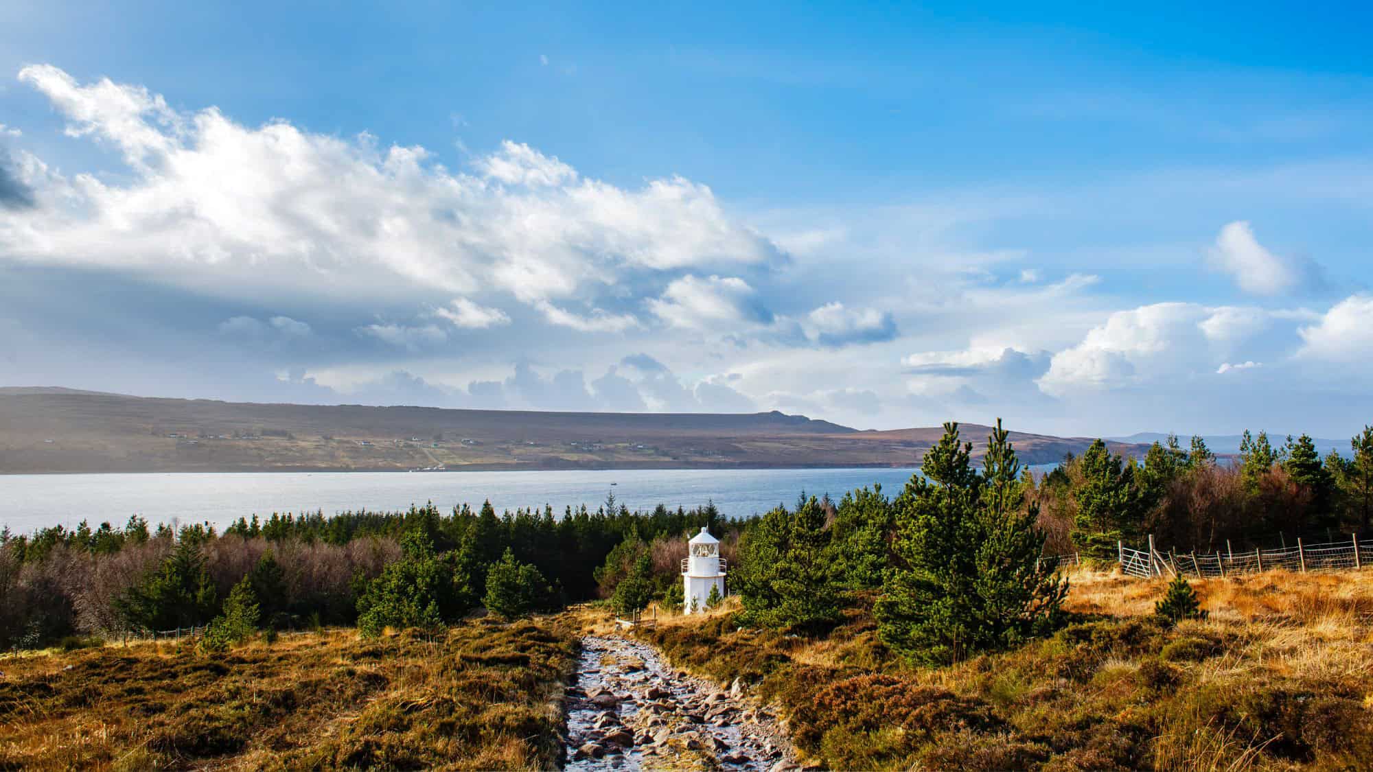 A secluded white lighthouse tucked among moorland and pine trees on the edge of a calm loch, with a misty view of hills in the background.
