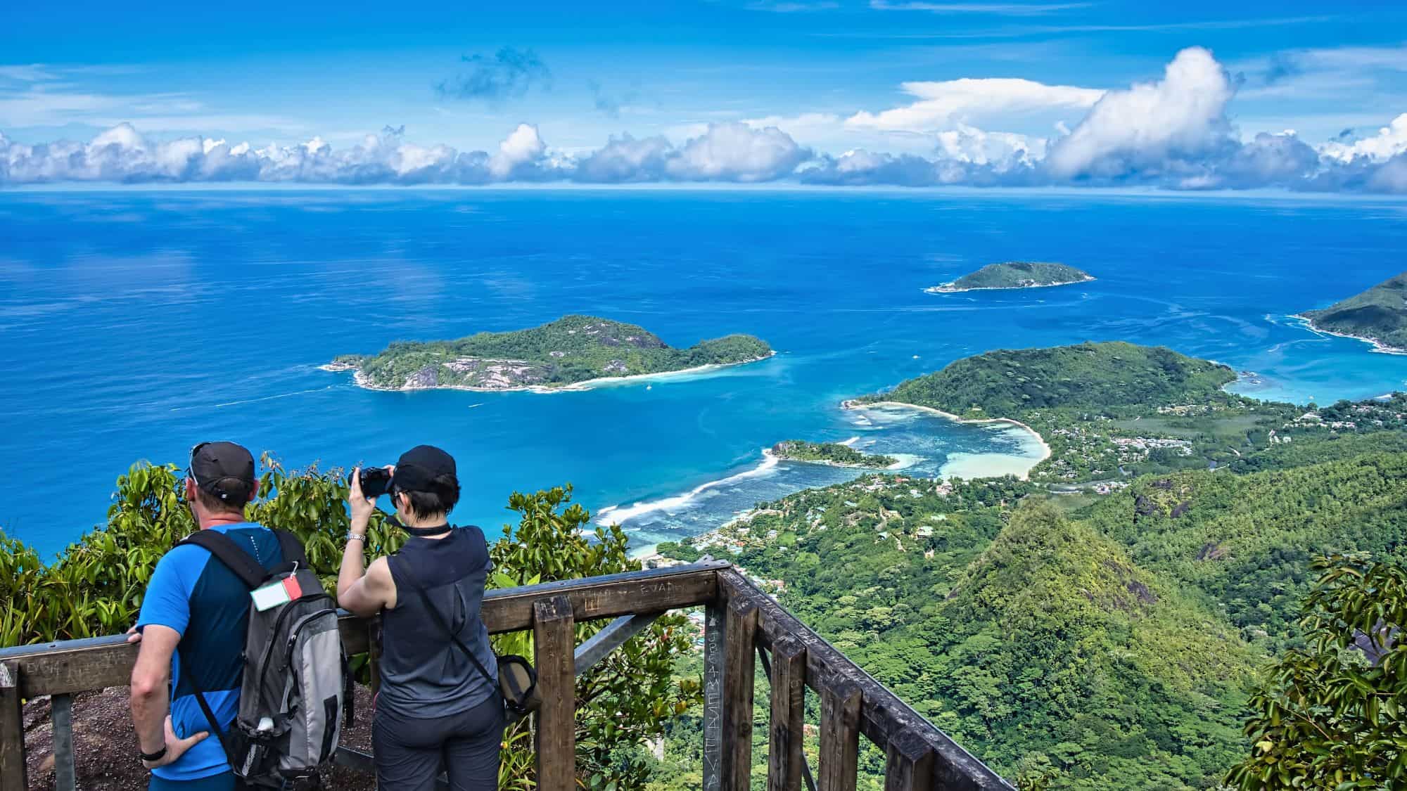 Two hikers admire a sweeping panoramic view of Seychelles’ coastline and lush islands from a wooden lookout deck atop Morne Blanc mountain trail.