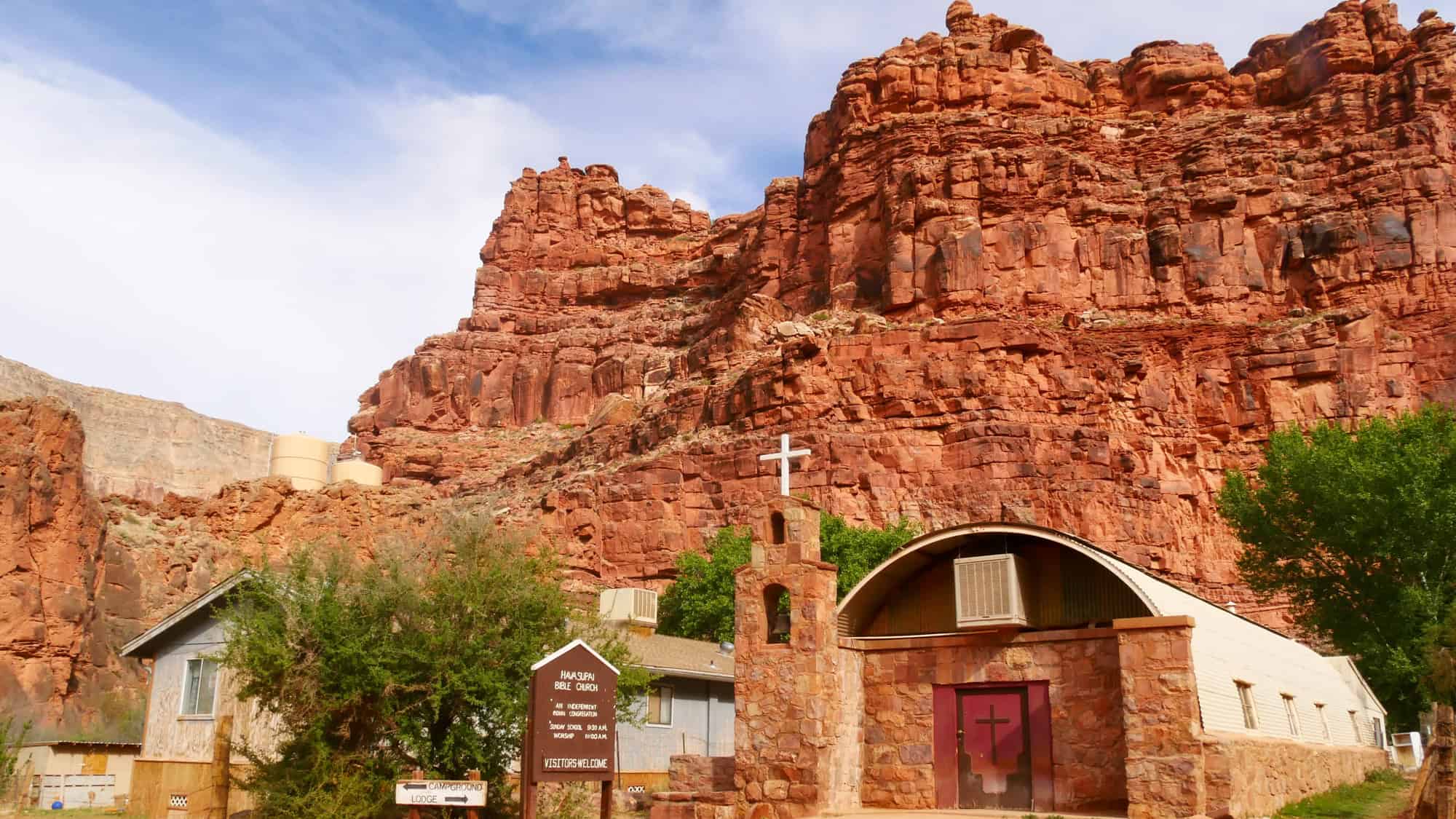 A church made of stone and concrete nestled beneath dramatic red rock cliffs in the remote desert village of Supai; a sign in front welcomes visitors.