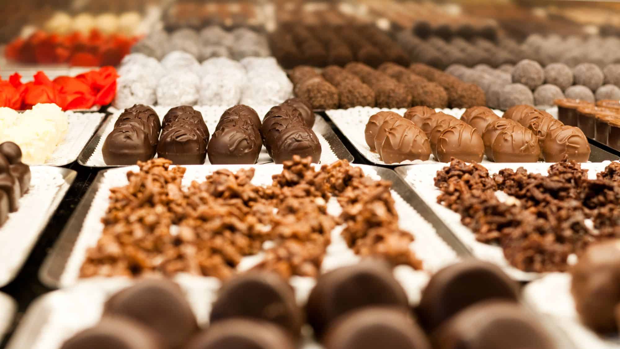 Close-up of a decadent assortment of Swiss chocolates arranged on trays, including white, milk, and dark truffles, with some topped in cocoa powder and others shaped into clusters or glossy domes.

