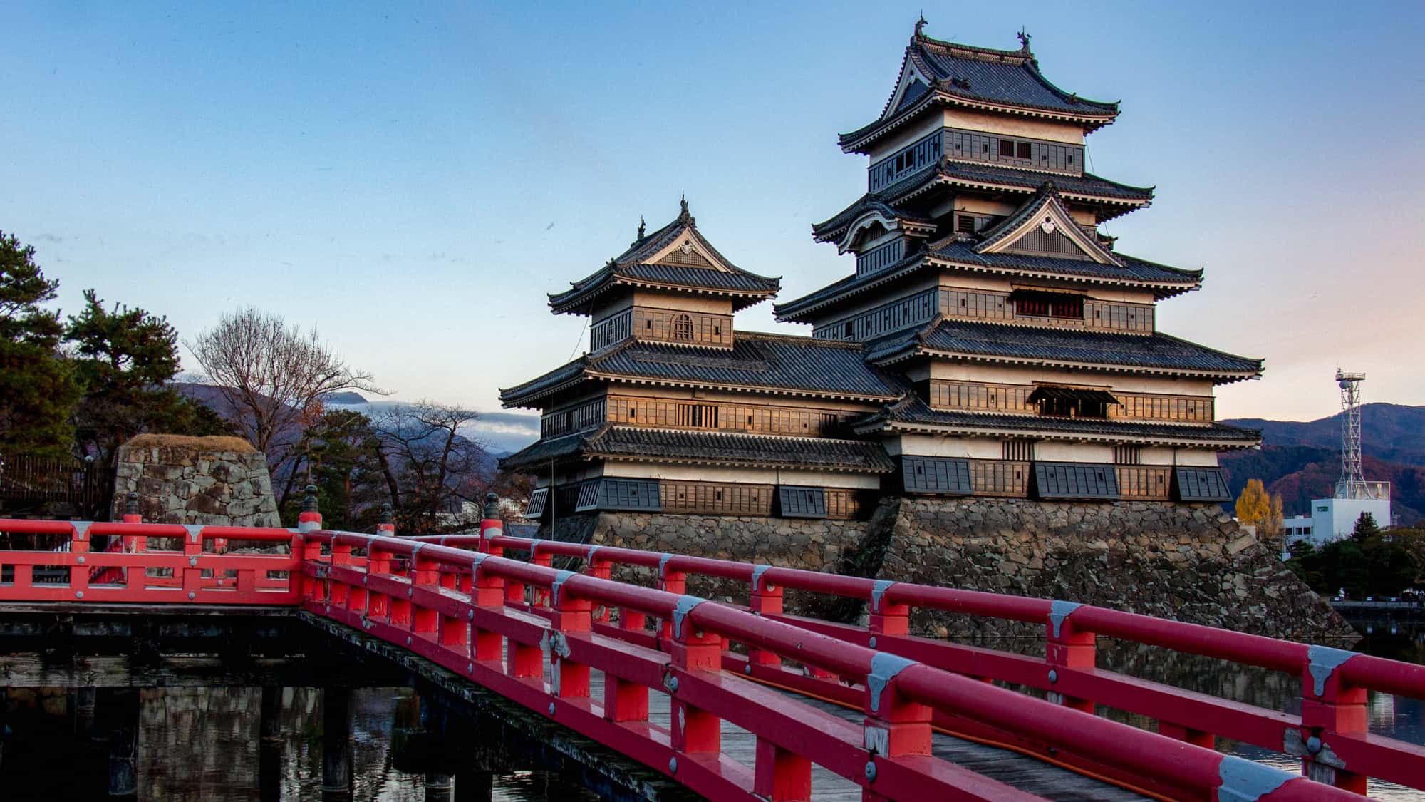 Traditional Japanese architecture at Matsumoto Castle with its dark wooden tiers, stone base, and a red pedestrian bridge spanning the water at sunset in Takayama, Japan.