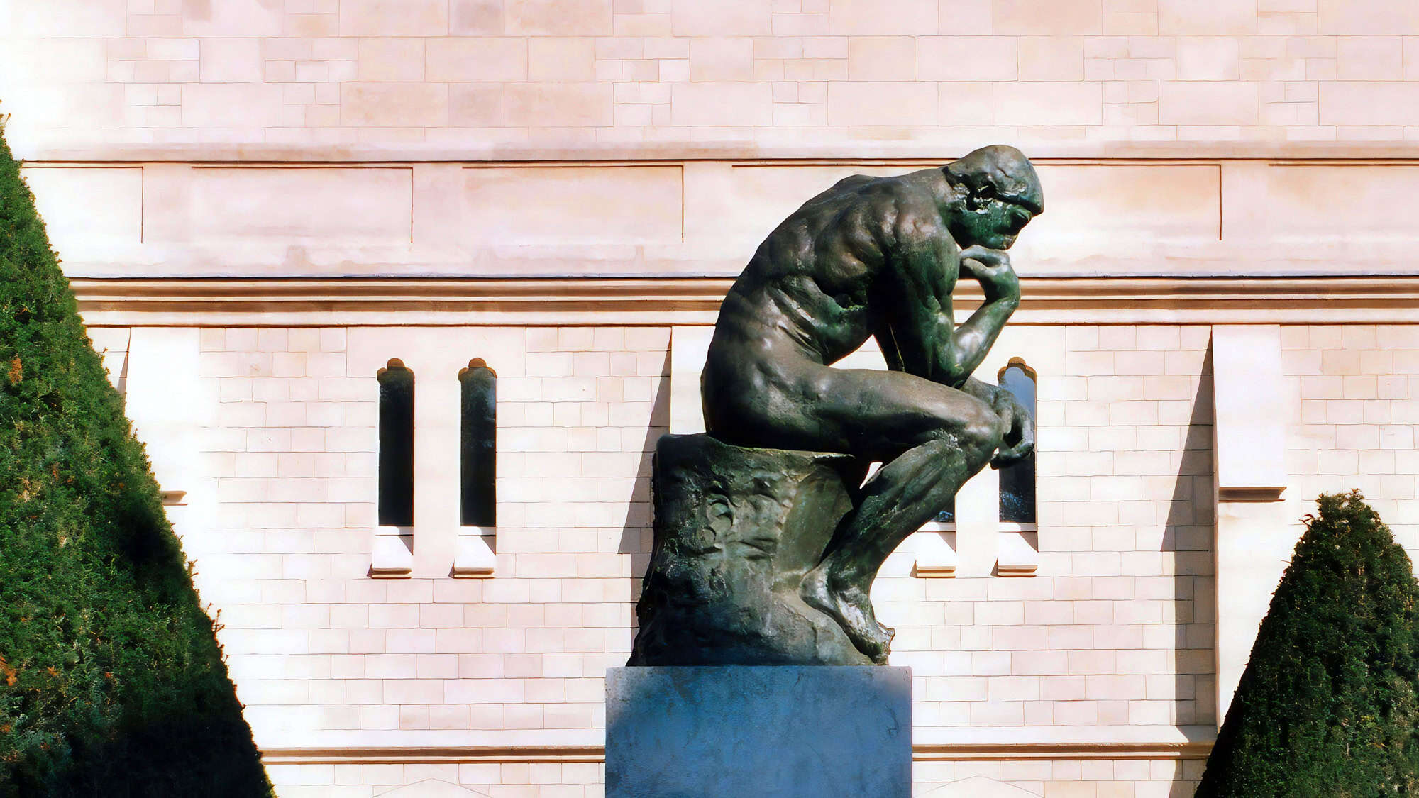 A bronze sculpture of a muscular man seated with his chin resting on one hand, deep in contemplation, placed in front of a light stone building. The figure, famously known as The Thinker by Auguste Rodin, is framed by manicured greenery.