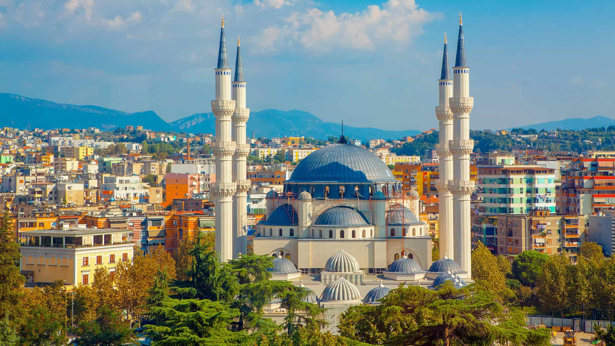 A striking image of Tirana’s skyline centered on the modern Grand Mosque with its four towering minarets and large blue dome, surrounded by colorful apartment buildings and backdropped by hazy mountains.