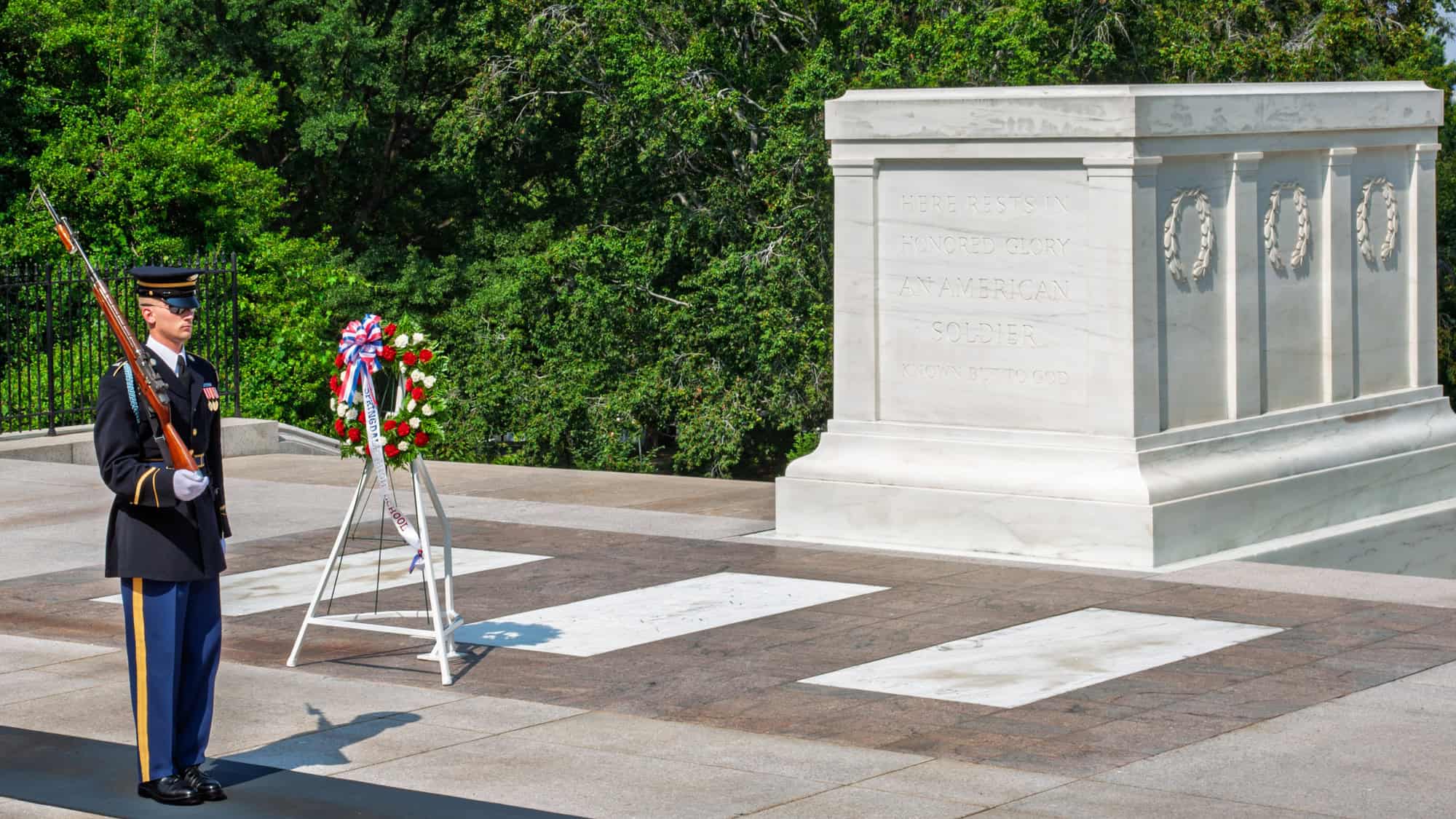 A uniformed guard stands in solemn vigil beside the Tomb of the Unknown Soldier at Arlington National Cemetery. The marble tomb bears the inscription, “Here rests in honored glory an American soldier known but to God,” beside a floral wreath.