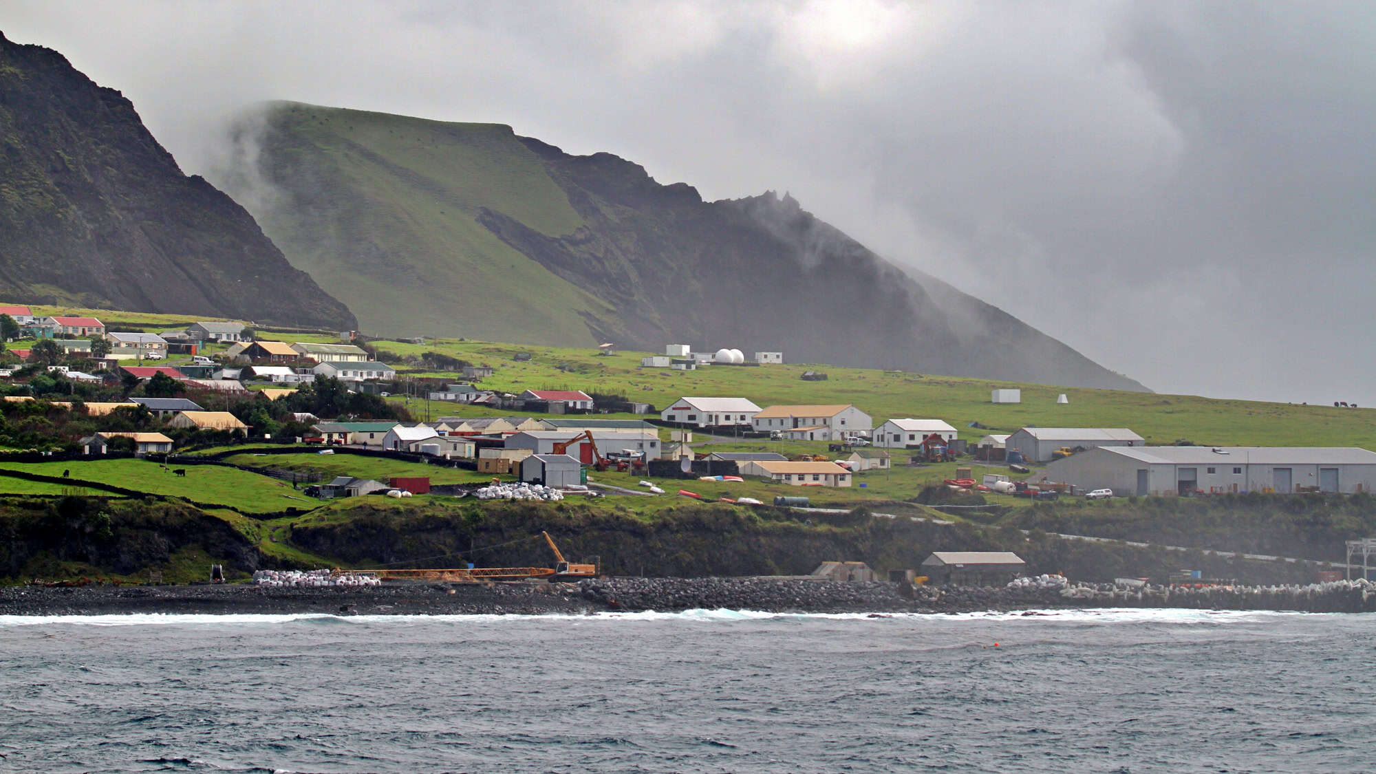 A windswept volcanic island settlement with low houses and green fields bordered by cliffs and shrouded in coastal mist.