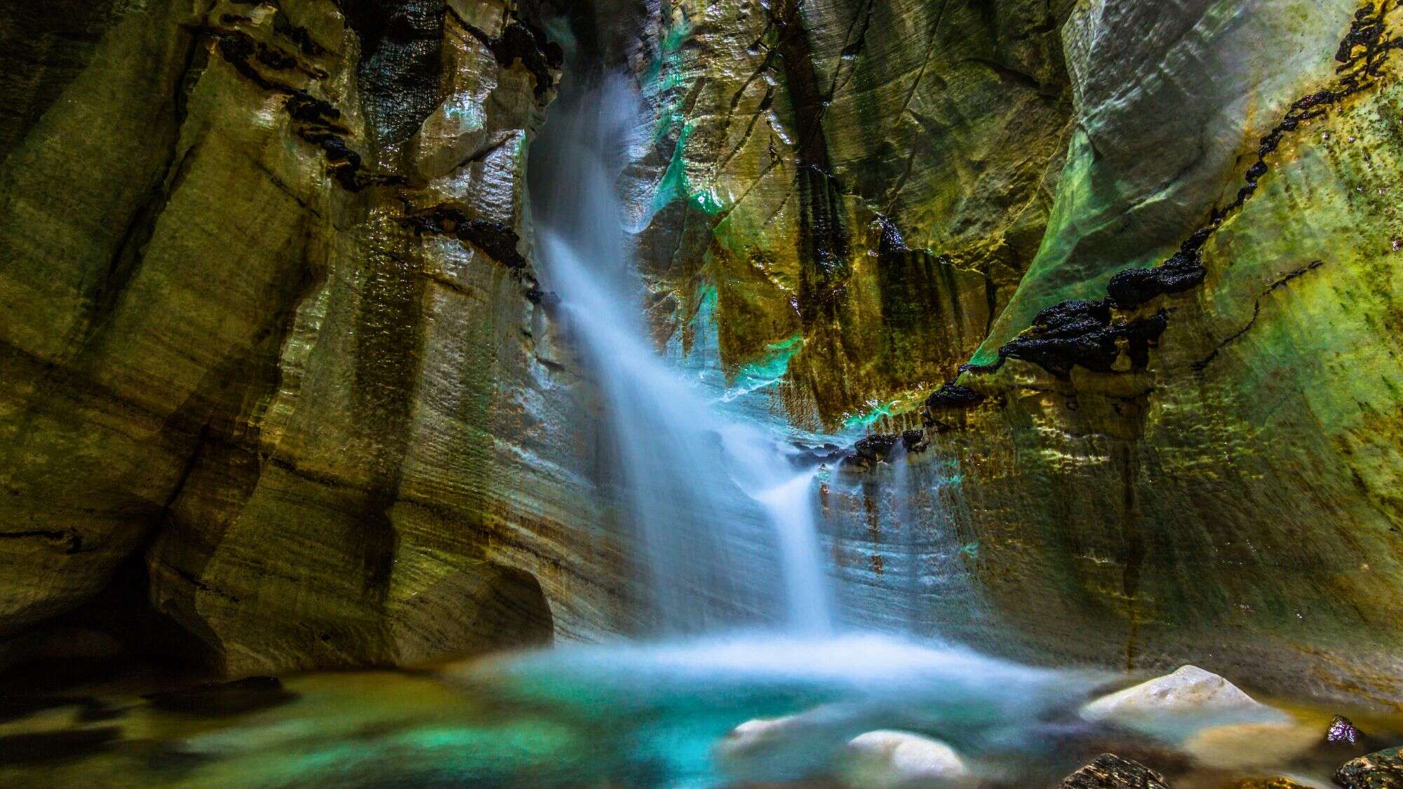 A narrow waterfall cascades through a mossy, jagged cave wall in Norway’s Trollkirka cave system, with turquoise water pooling below and light glinting off the wet rock surfaces.