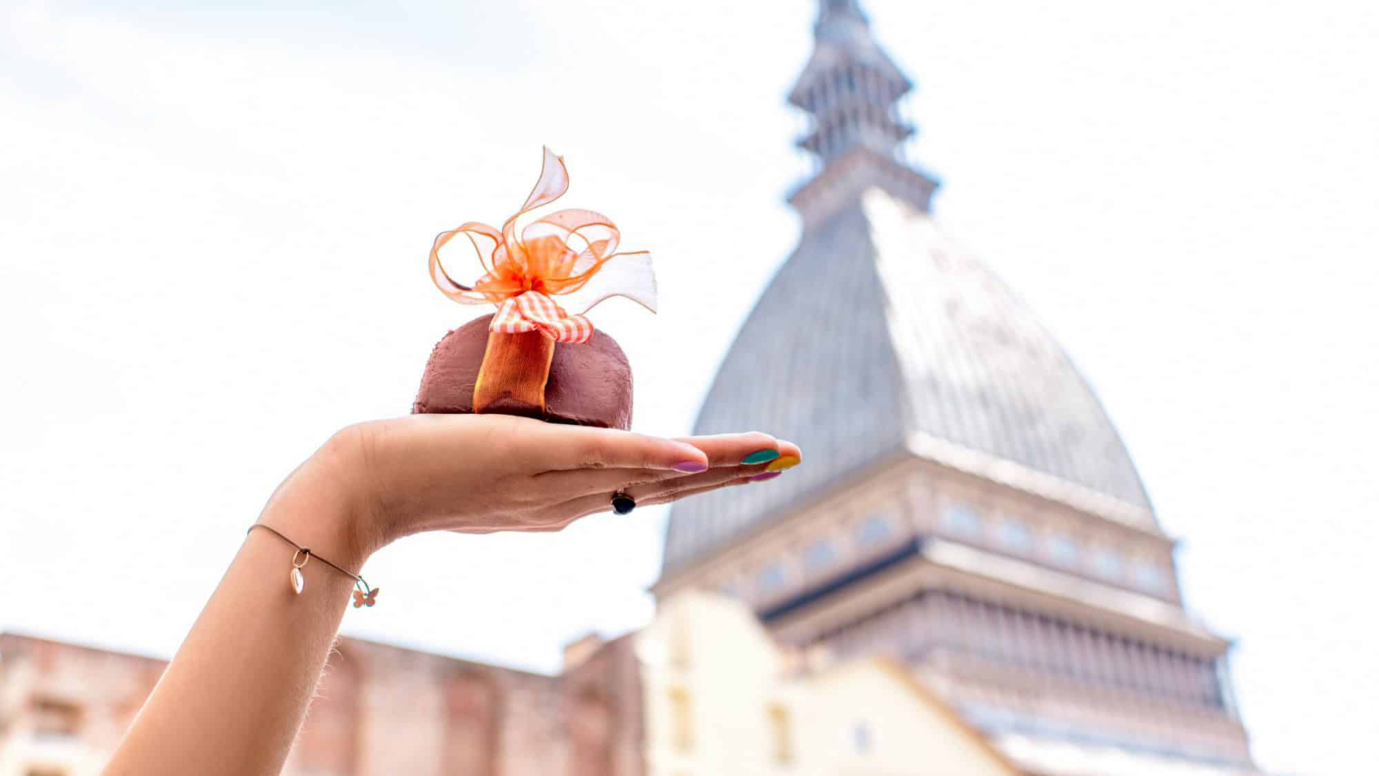 A hand with rainbow-painted nails holds a chocolate dessert tied with a festive orange bow, with Turin’s Mole Antonelliana softly blurred in the background for a charming Italian touch.