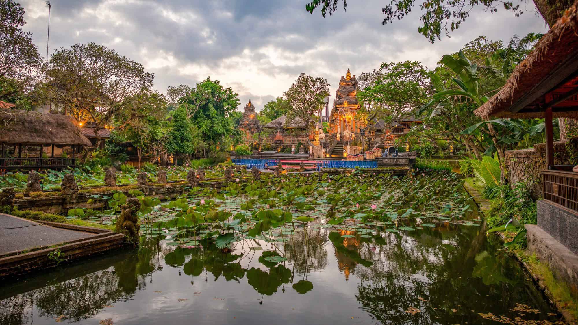 Tranquil view of a Balinese temple surrounded by a blooming lotus pond, lush greenery, and traditional architecture at dusk in Ubud, Bali.