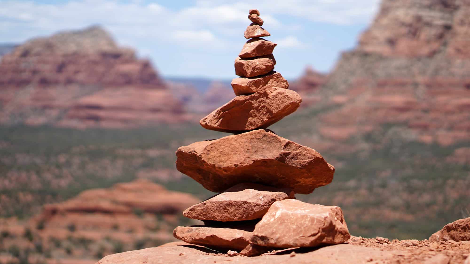A carefully stacked tower of red sandstone rocks forms a balanced cairn against the dramatic, blurred backdrop of Sedona's iconic red rock formations and blue sky.