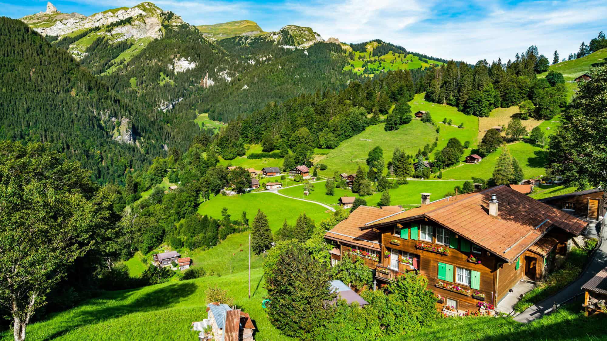 A charming Swiss alpine village with wooden chalets, vibrant green meadows, and forested mountains in the background under a bright summer sky.