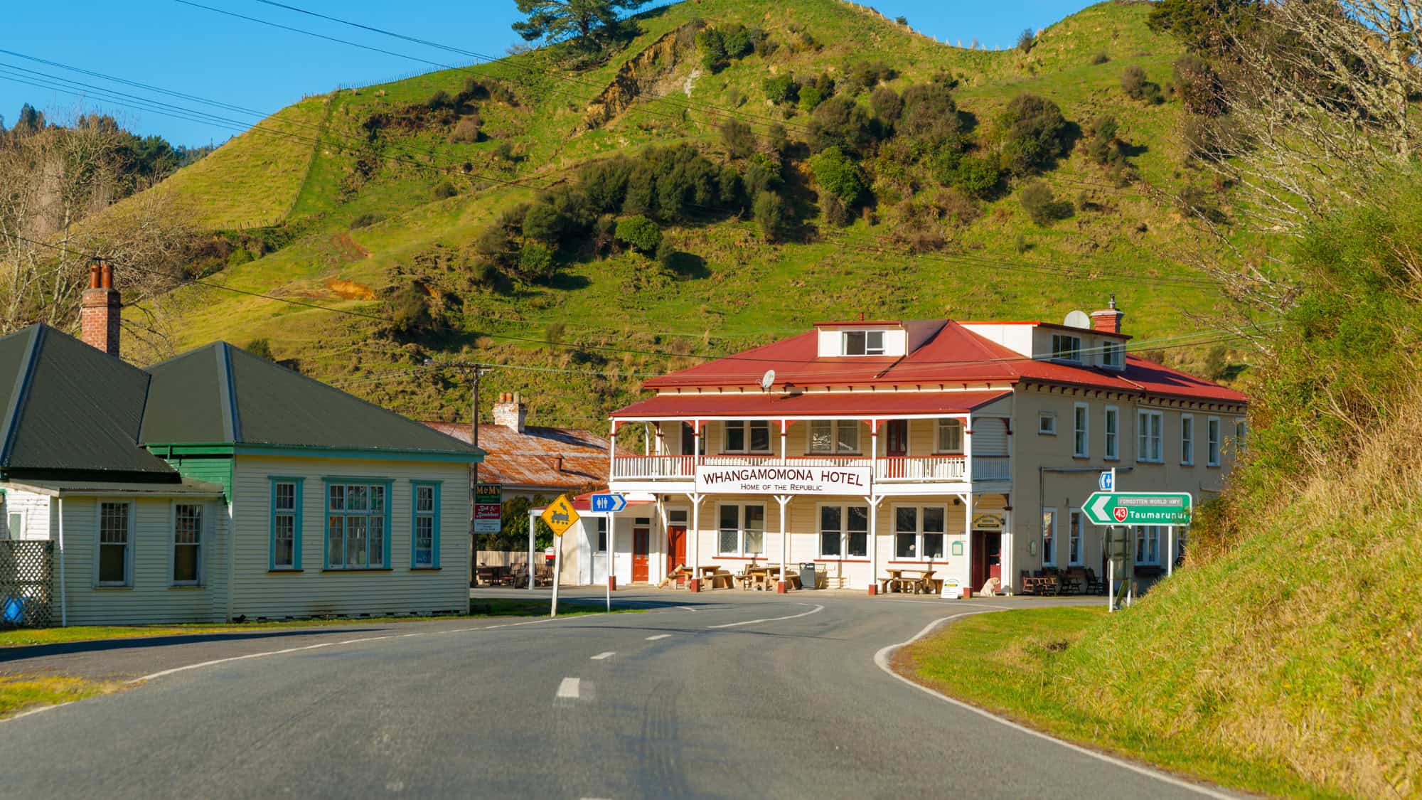 A rural roadside scene featuring the Whangamomona Hotel with a sign declaring it the “Home of the Republic,” flanked by green hills and modest buildings under a clear blue sky.