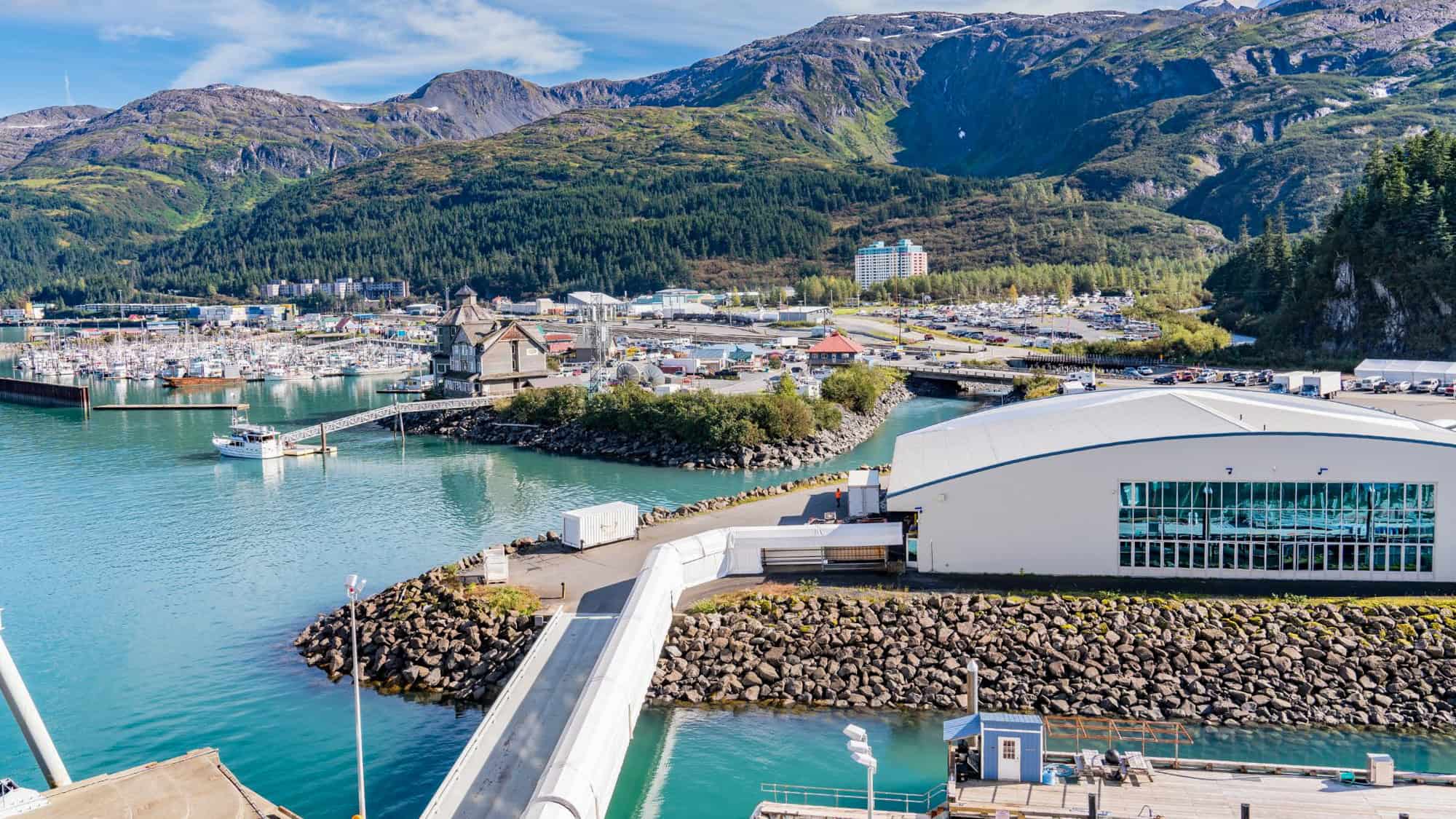 A coastal Alaskan town with a busy harbor, surrounded by forested mountains and rugged peaks; the town’s unique feature is its centralized high-rise residential building visible in the distance.