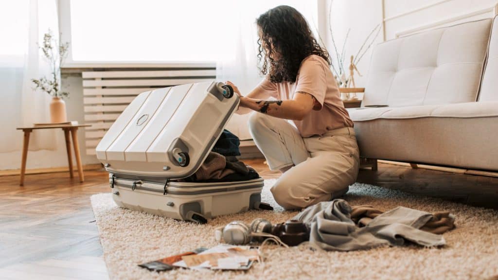 A woman kneels on the floor of a cozy living room, closing a hard-shell suitcase with clothes, headphones, and travel brochures scattered around, preparing for a trip.