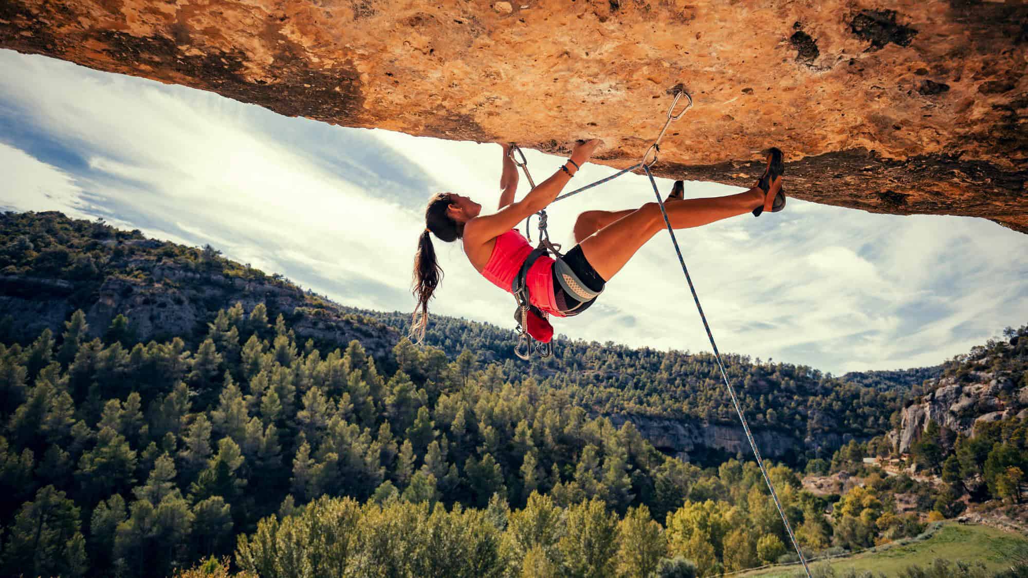 A woman in a red tank top and climbing gear scales an overhanging rock face, suspended high above a forested canyon landscape.