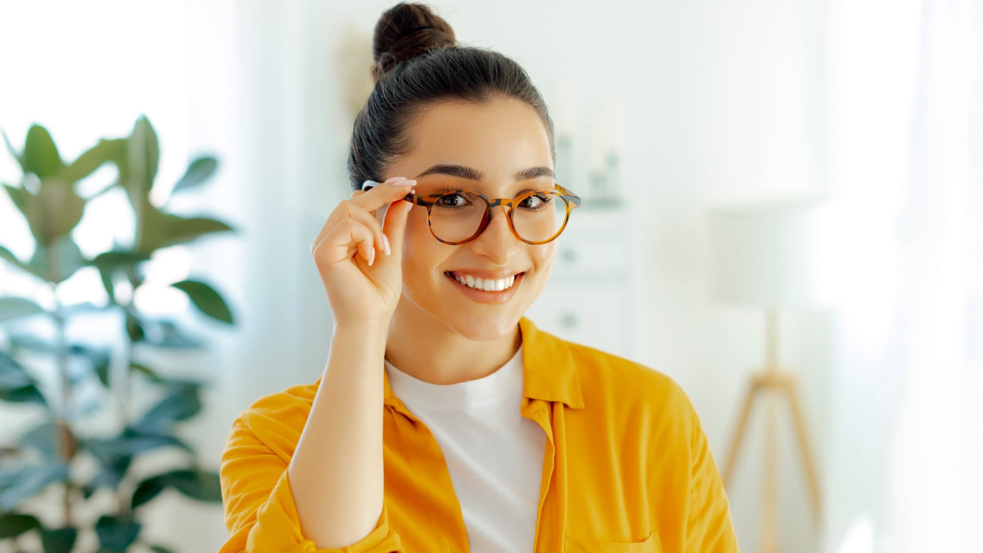 A cheerful woman in a yellow shirt and glasses smiles warmly at the camera, standing in a brightly lit room with soft white and green décor.