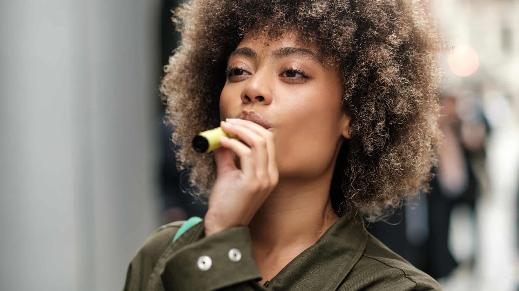A woman with natural curly hair and a green jacket holds a yellow e-cigarette to her lips, looking off into the distance with a thoughtful expression.