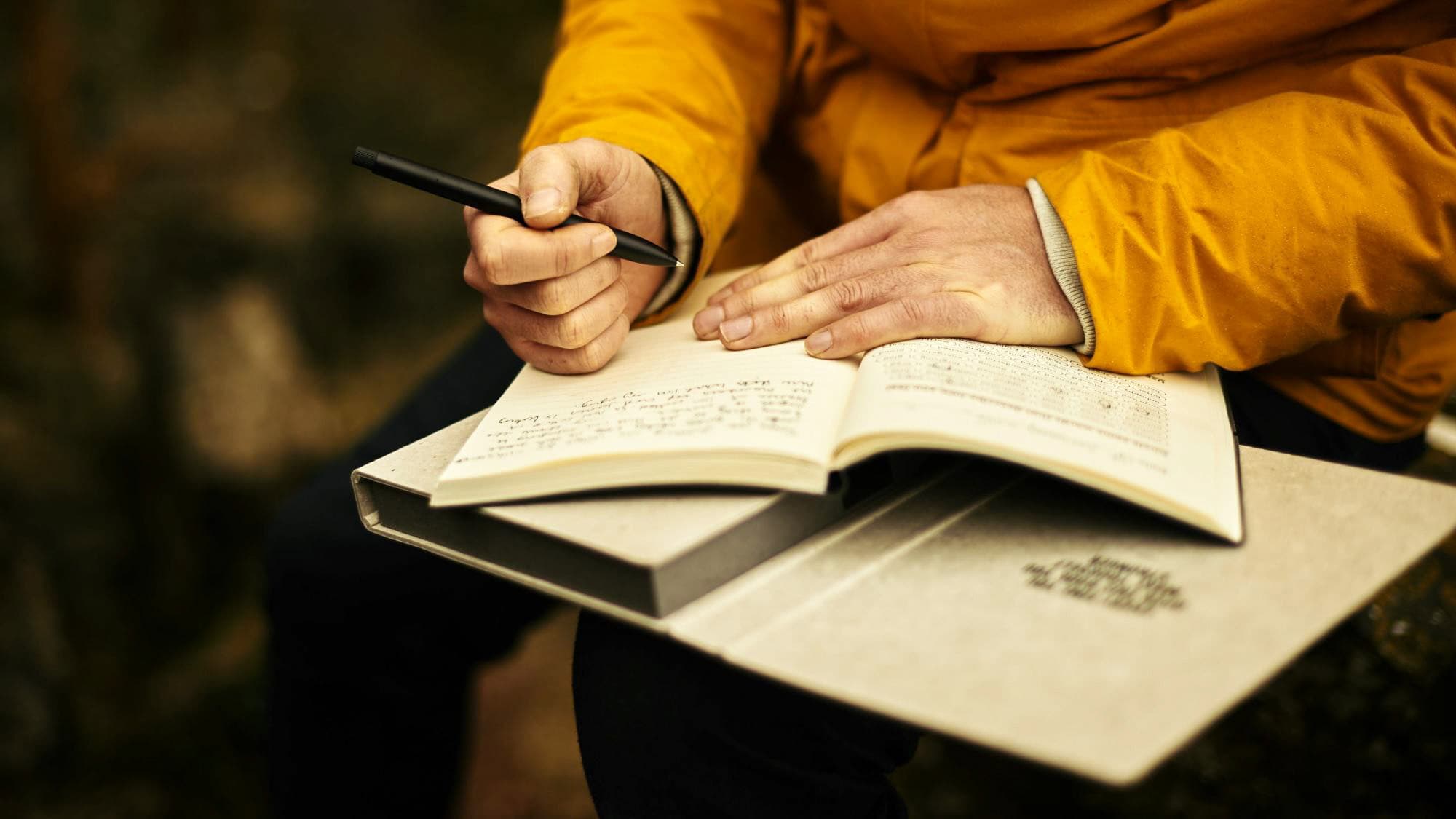 A close-up of a person in a yellow jacket writing in a journal with neat handwriting while seated outdoors, with another book or notebook resting on their lap.