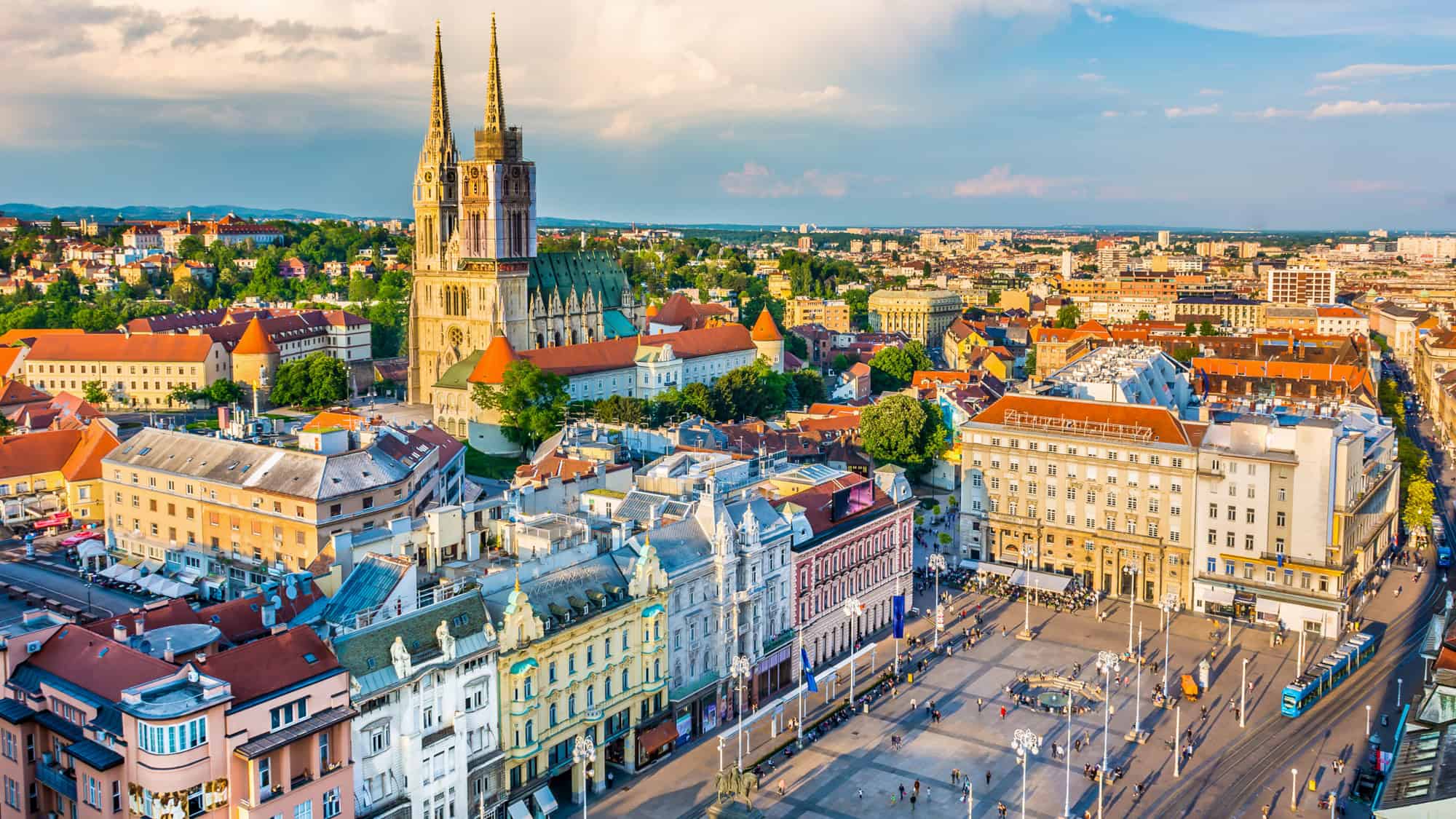 A panoramic view of Zagreb showcasing the twin spires of the iconic Zagreb Cathedral rising above a blend of red-roofed buildings, historic architecture, and modern city sprawl, all bathed in warm evening light.