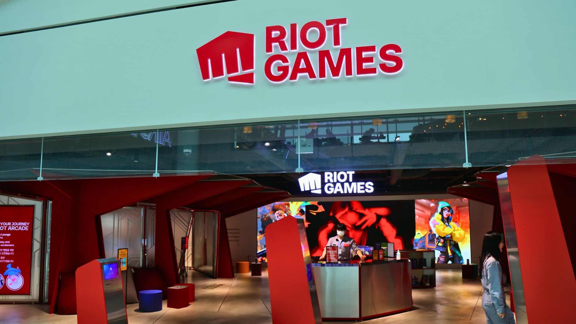 A sleek, futuristic Riot Games-themed arcade lounge with a bold red entrance, large digital screens, and gaming merchandise on display, located inside an airport terminal.