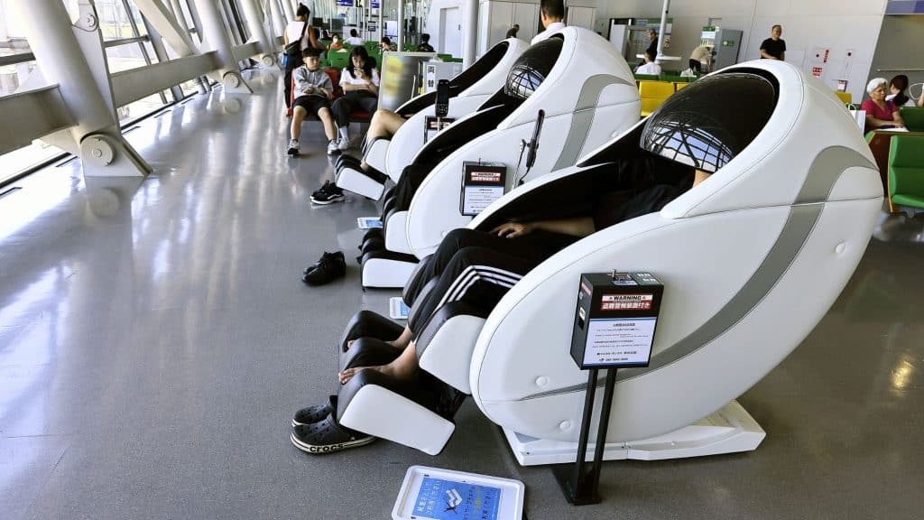 A row of high-tech white massage chairs with capsule-like design occupied by travelers, situated near large windows in a quiet section of an airport terminal.