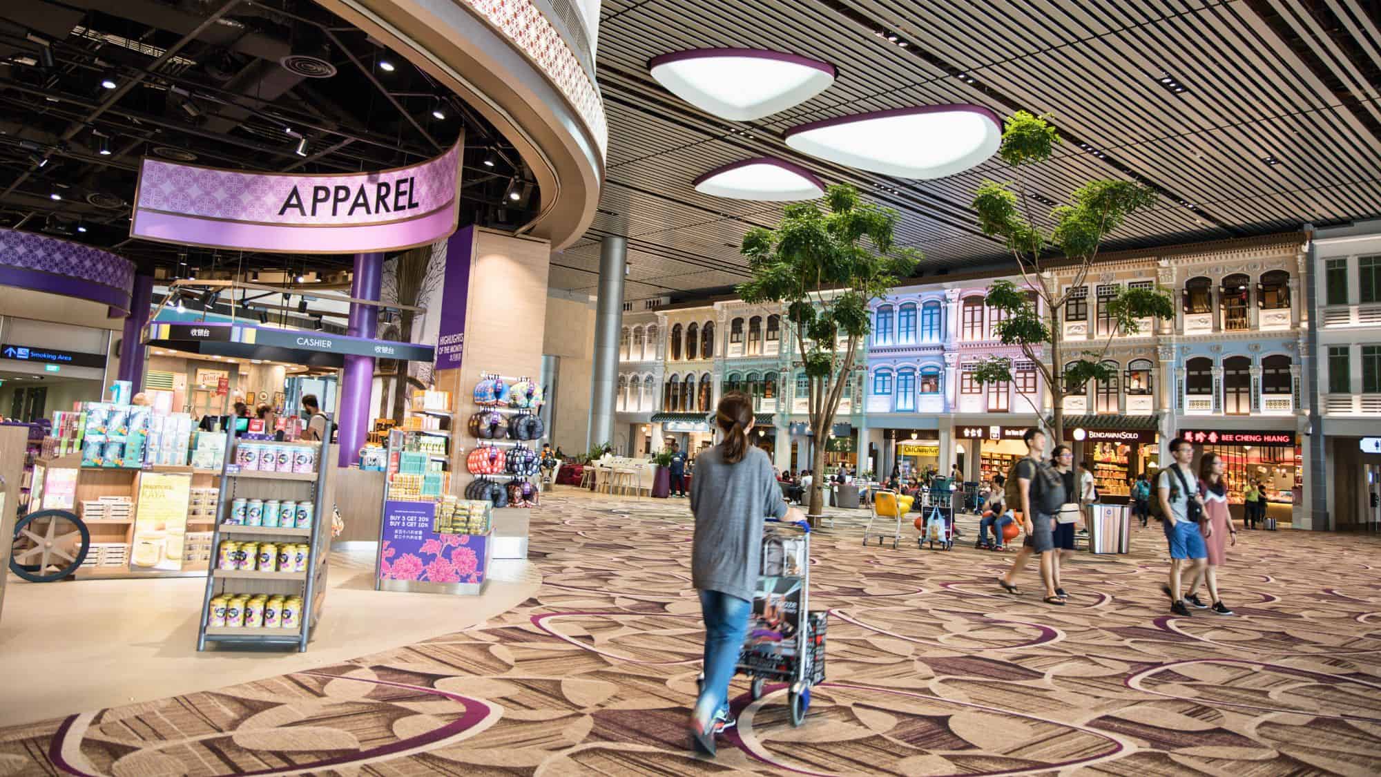 A vibrant airport shopping area with mock heritage-style building facades, colorful signage, and a variety of duty-free shops under dramatic modern lighting.
