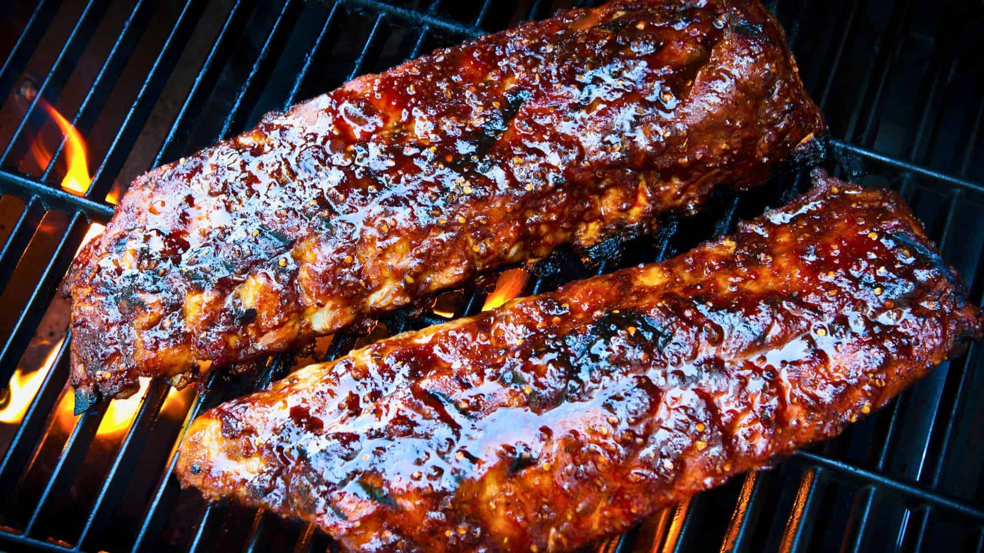 Two racks of glazed, saucy barbecue ribs sizzle over an open flame on a grill, their caramelized surfaces glistening with a smoky char.