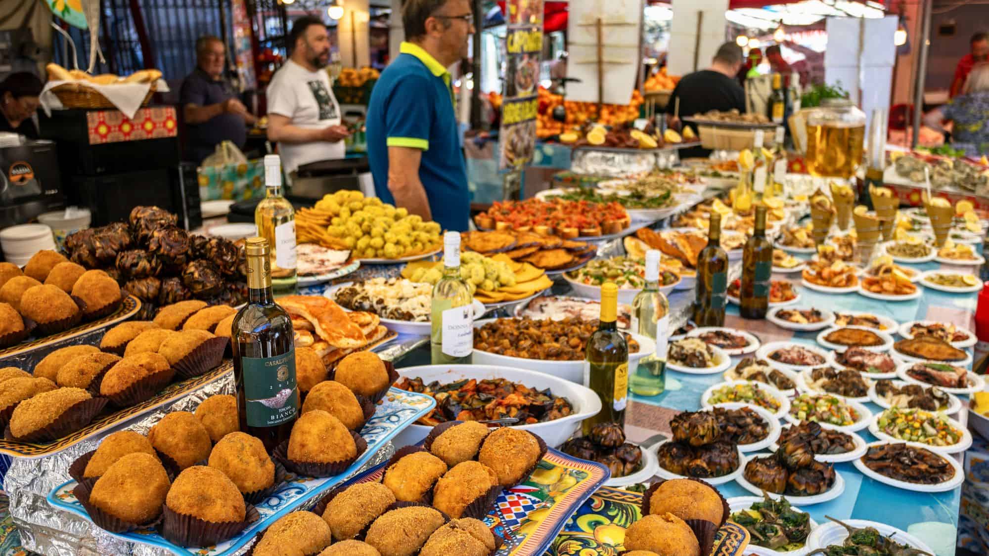 A colorful and abundant display of Sicilian street food including arancini, grilled vegetables, pasta dishes, and bottles of white wine on patterned ceramic plates at Ballarò Market in Palermo.