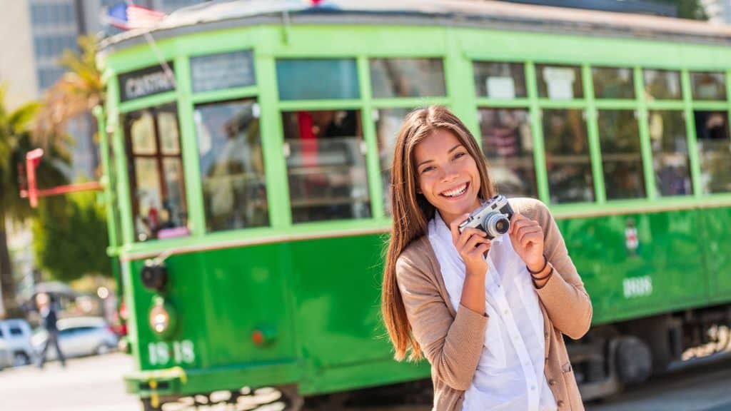 A smiling young woman poses with a vintage camera in front of a bright green historic streetcar with large windows and an American flag, suggesting a cheerful travel moment in an urban setting.