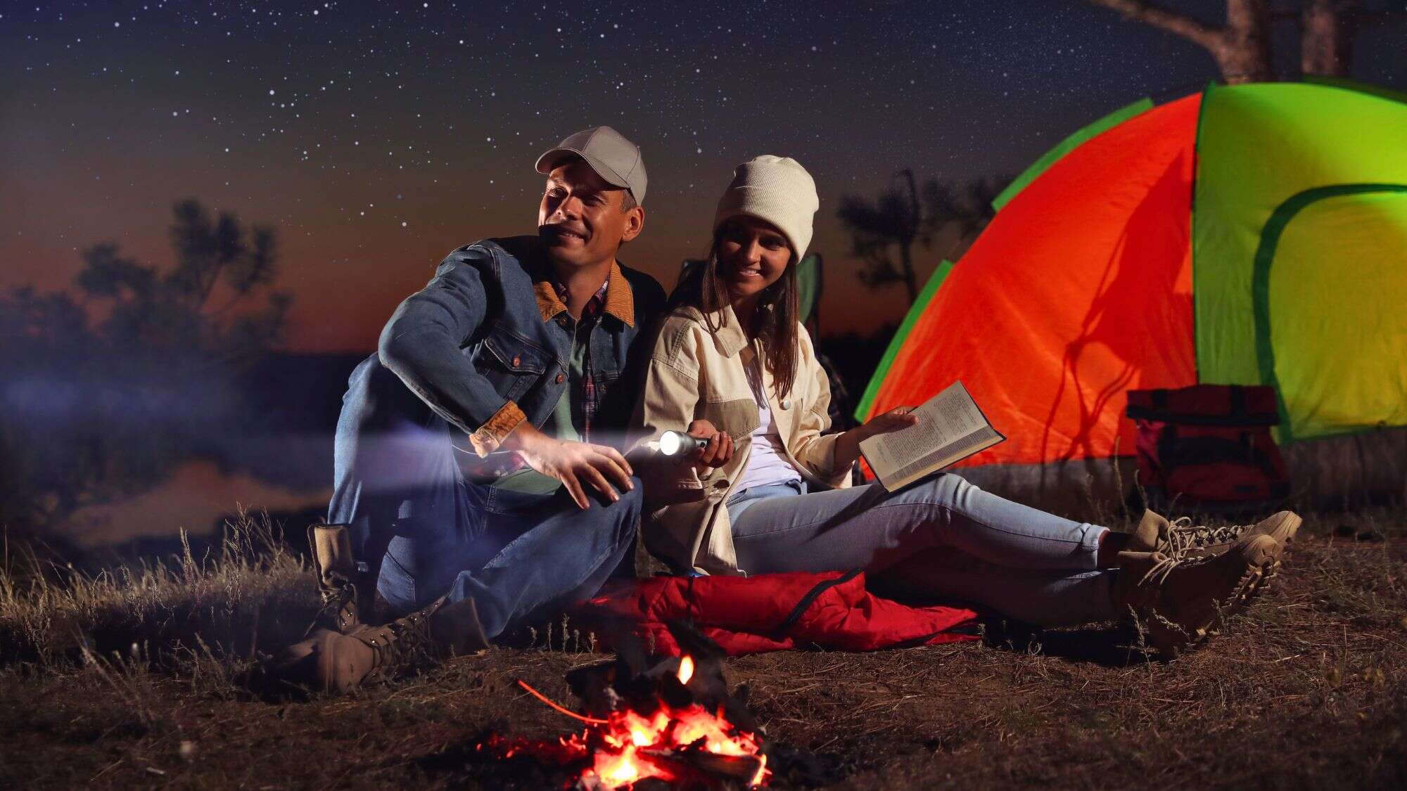 A couple sits by a glowing campfire under a starlit sky, with a colorful tent behind them; the man holds a flashlight while the woman reads a book, both dressed warmly in beanies and boots.