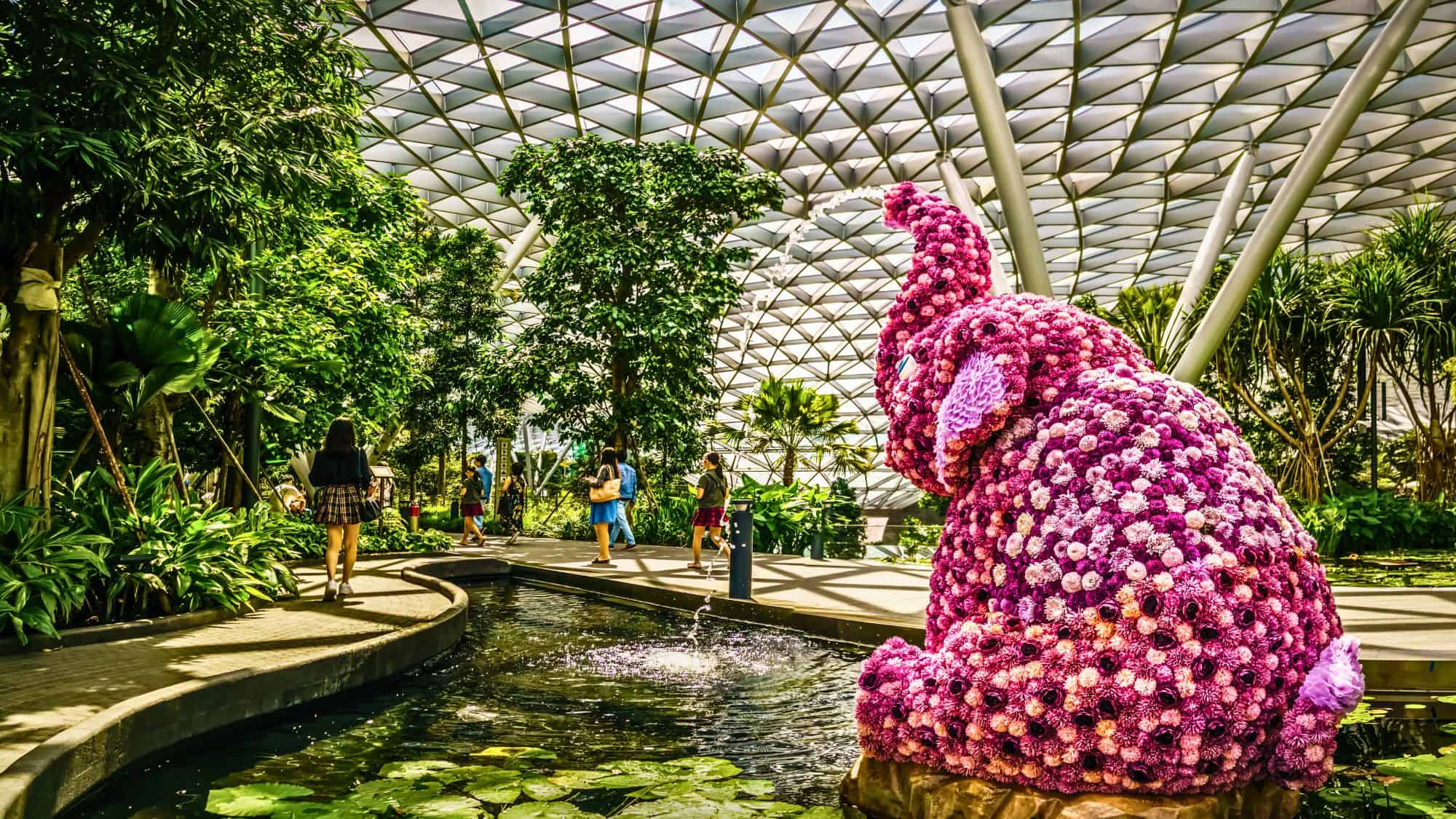 A vivid floral elephant sculpture made of pink and purple blooms sits beside a lily pond under the Jewel Changi Airport’s iconic glass dome, surrounded by lush indoor greenery and strolling visitors.
