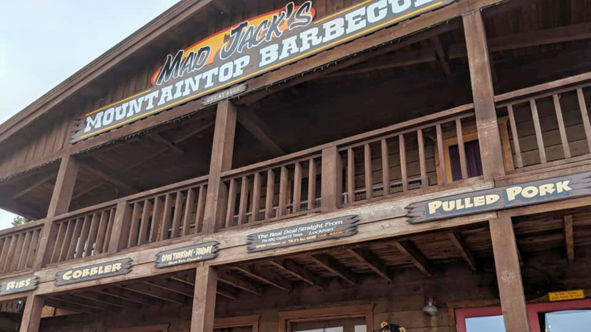 A rustic, wooden two-story restaurant front with signage reading "Mad Jack’s Mountaintop Barbecue" and menu highlights like ribs, cobbler, and pulled pork. The structure has a cabin-style charm with a balcony and a weathered look.