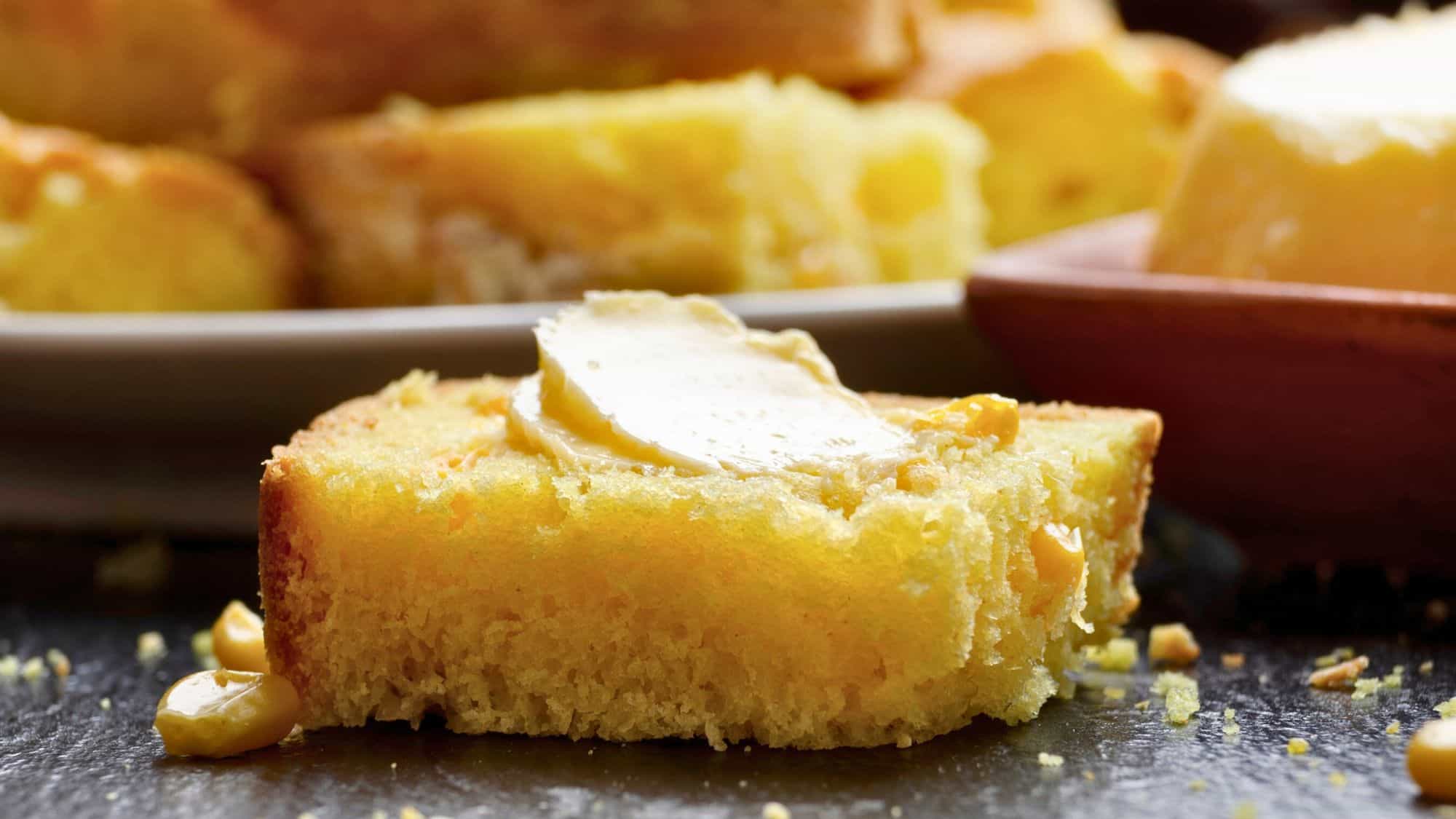 A slice of golden cornbread topped with a melting pat of butter sits on a dark surface, with visible kernels of corn and crumbs scattered nearby.