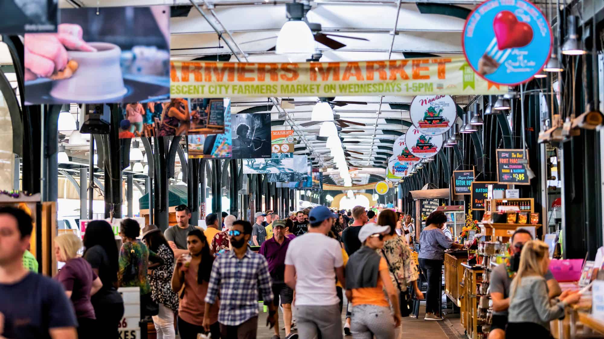 A lively indoor farmers market in New Orleans filled with shoppers exploring stalls selling food, drinks, and crafts, with overhead banners advertising local products and events.