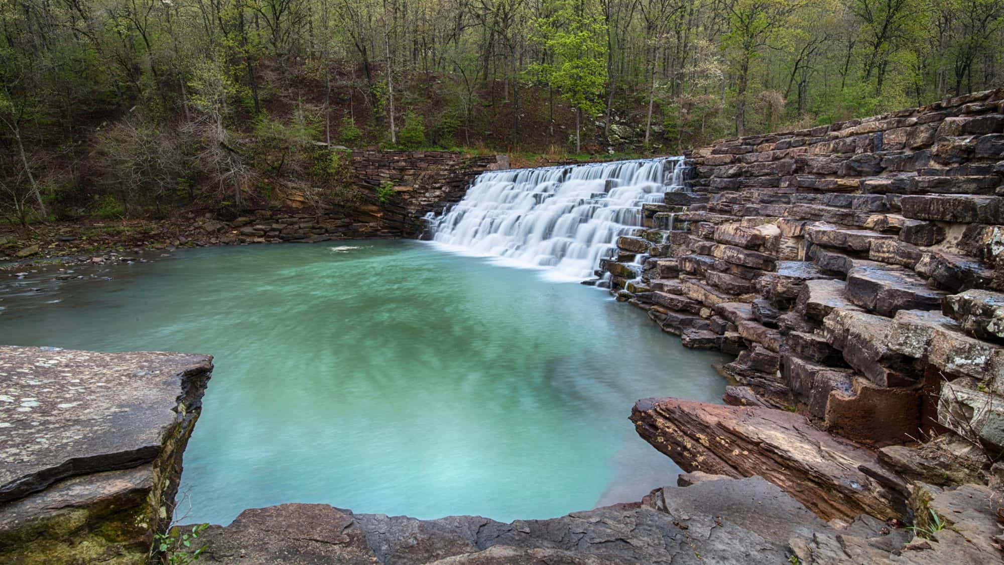 A waterfall cascades in a wide curtain over rocky ledges into a turquoise pool, surrounded by spring greenery at Devil’s Den State Park.