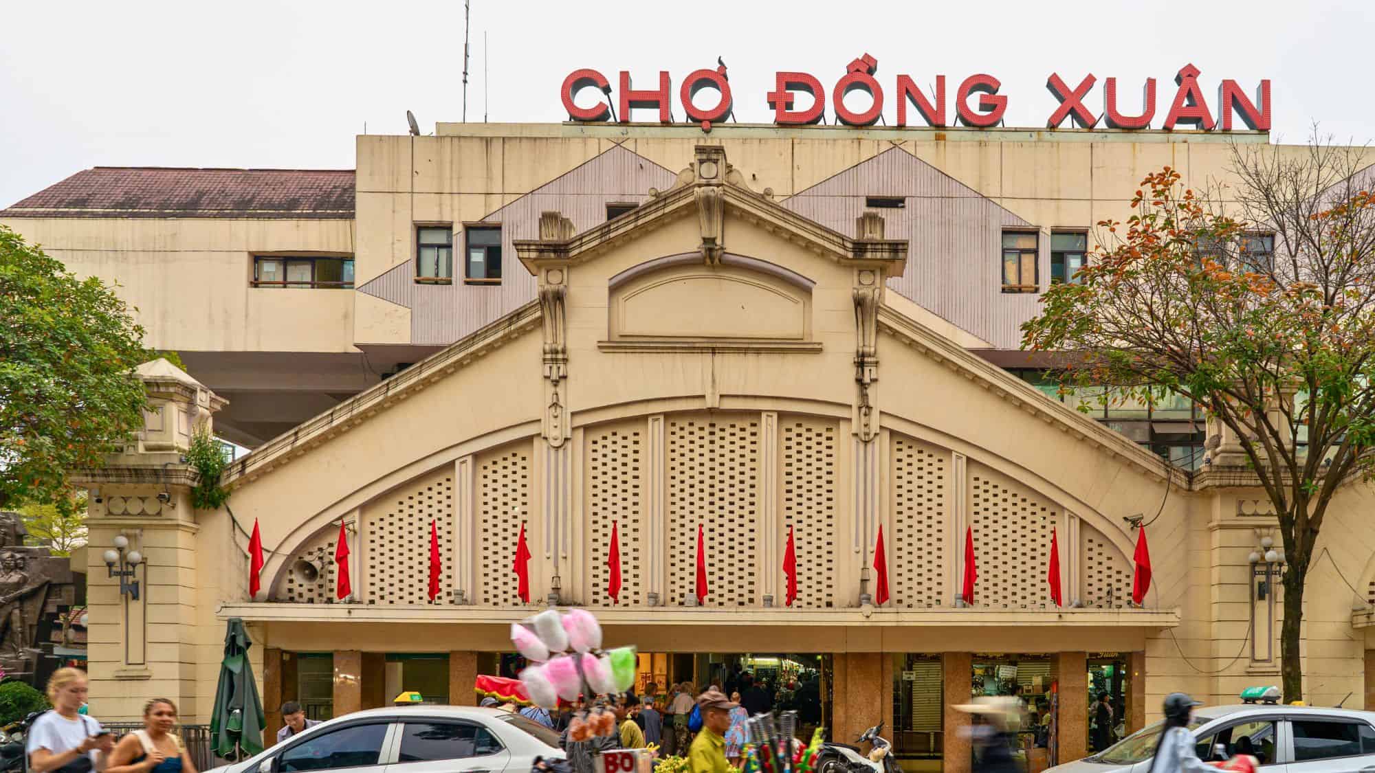 Street view of the entrance to Dong Xuan Market in Hanoi, Vietnam, featuring the market’s sign in bold red letters and shoppers walking in and out, with cotton candy and street vendors visible out front.