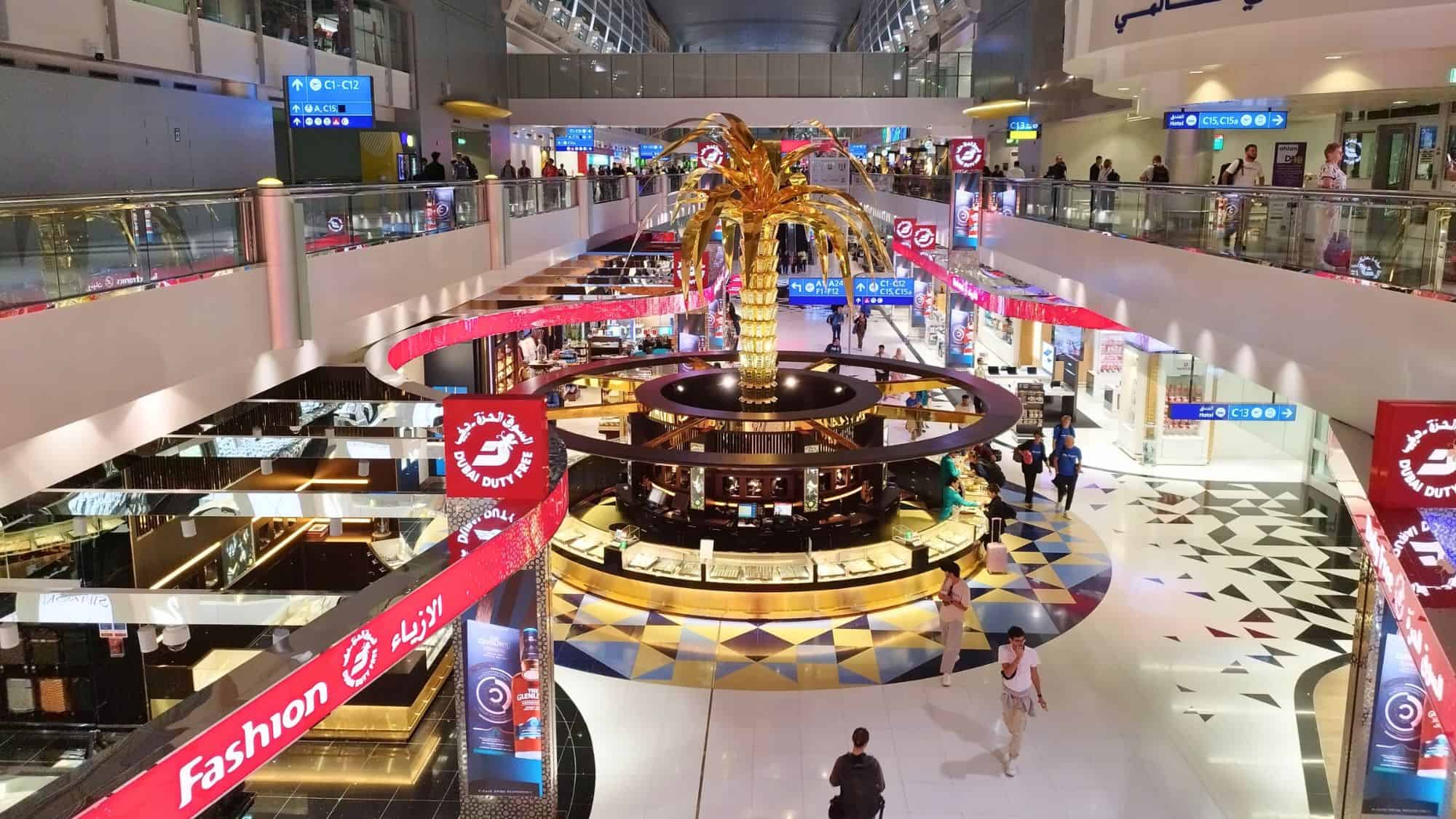 Dubai International Airport’s duty-free area showcases a giant golden palm tree at the center of luxury shopping surrounded by gleaming floors, red signage, and shoppers exploring multi-level boutiques.