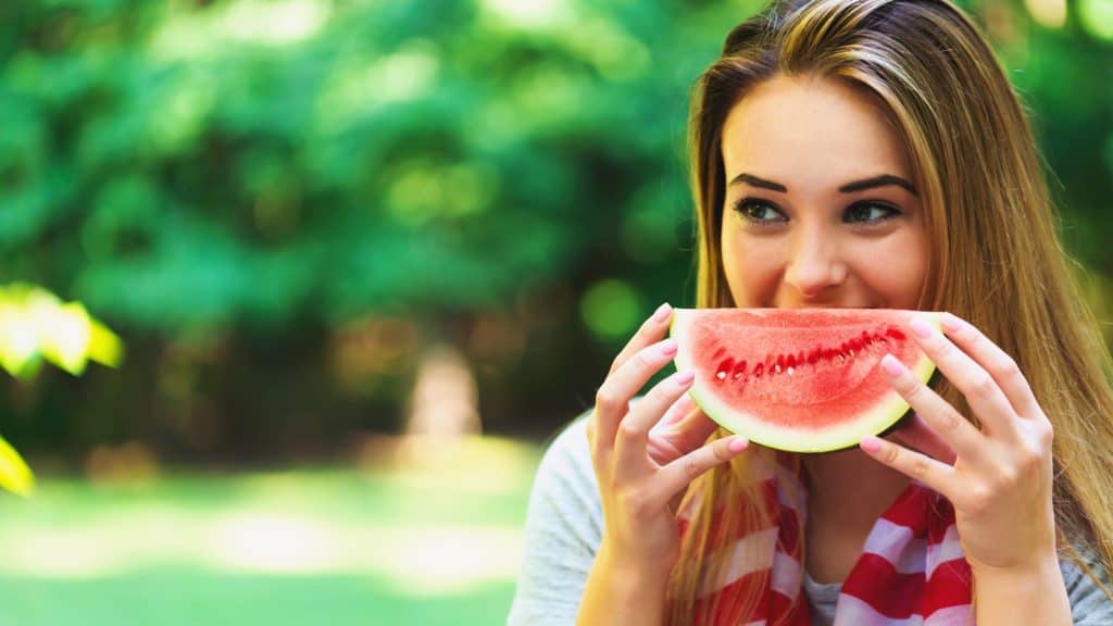 A young woman with long, light brown hair smiles while holding a large slice of watermelon in front of her face, set against a vibrant green, blurred outdoor background.