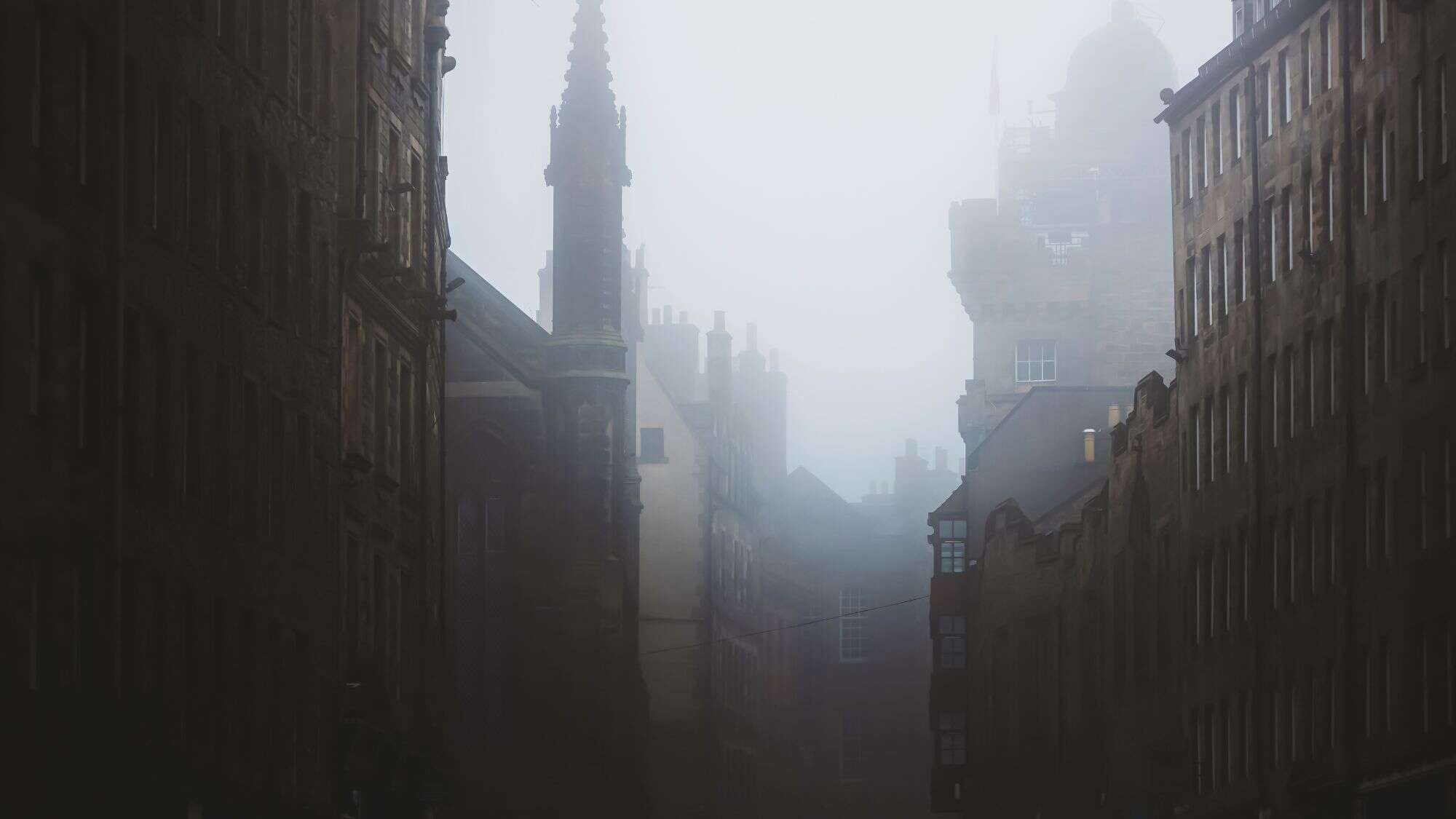 A moody, fog-filled street scene in Edinburgh where historic stone buildings and gothic spires fade into thick mist, evoking a ghostly, atmospheric vibe.