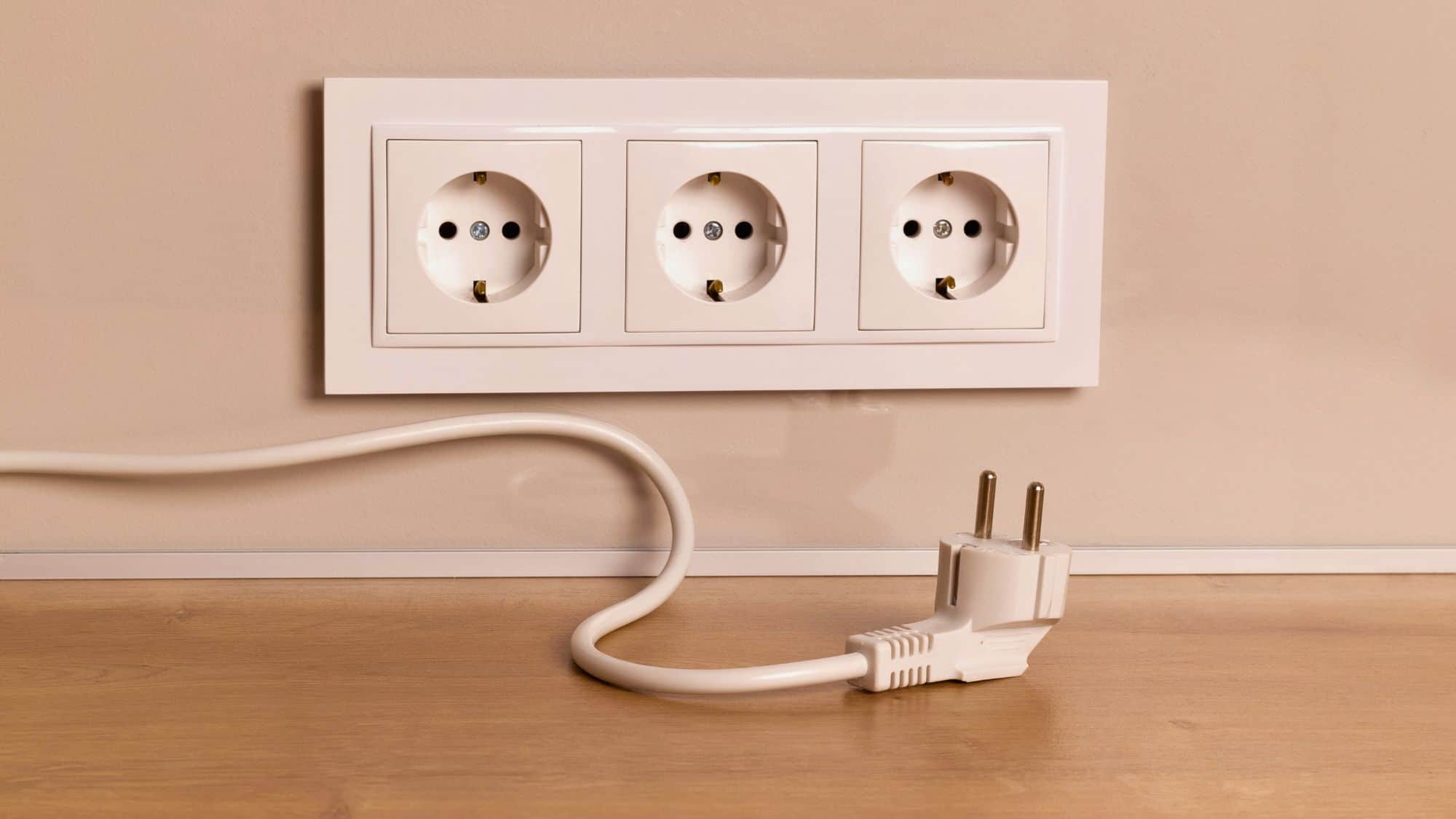 Three European-style electrical outlets mounted on a beige wall above a wooden surface, with a white power cord and plug placed in front.