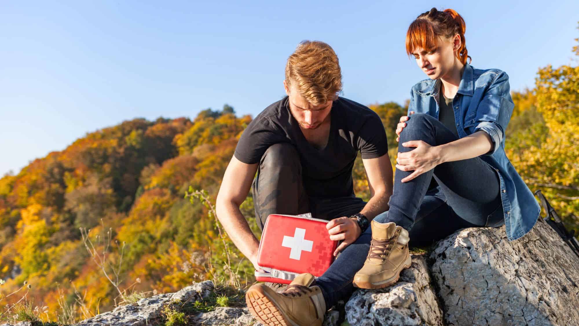 A hiker uses a red first aid kit to tend to his companion’s injured leg while sitting on a rocky outcrop, surrounded by autumn-colored trees.