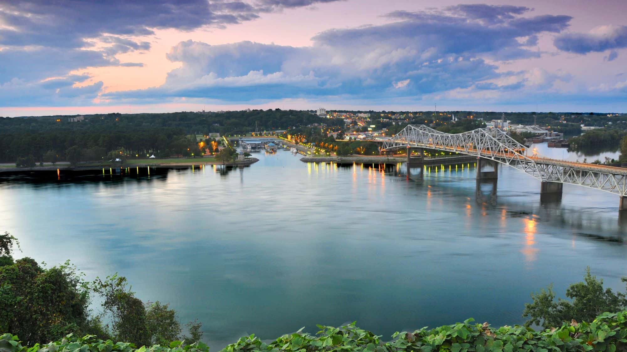 Scenic twilight view of Florence, Alabama featuring a calm river, a long truss bridge, and glowing city lights reflecting on the water under a colorful sunset sky.