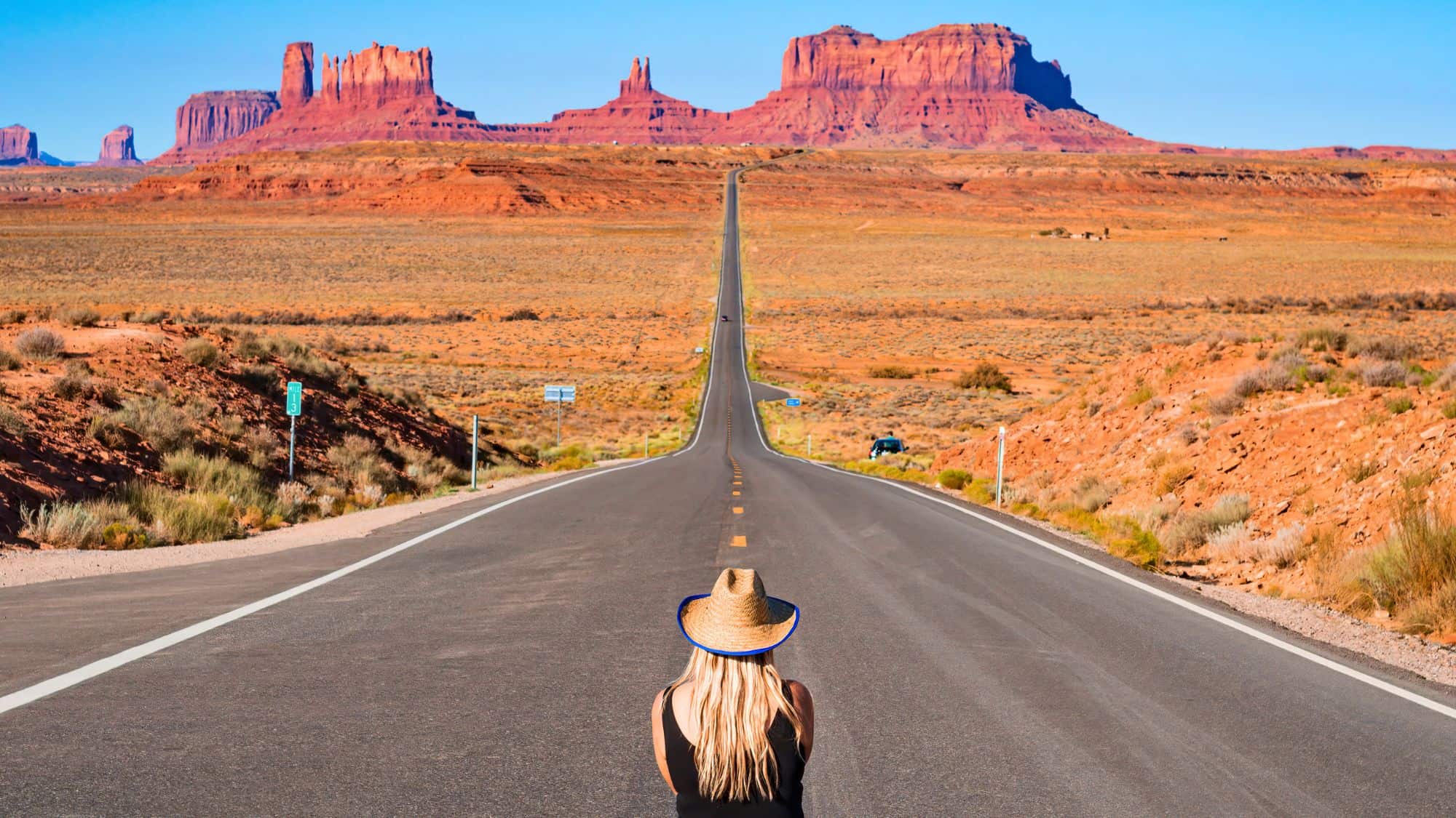 A woman wearing a straw hat sits in the middle of a long, empty desert highway that stretches straight toward towering red rock formations in Monument Valley under a clear blue sky.