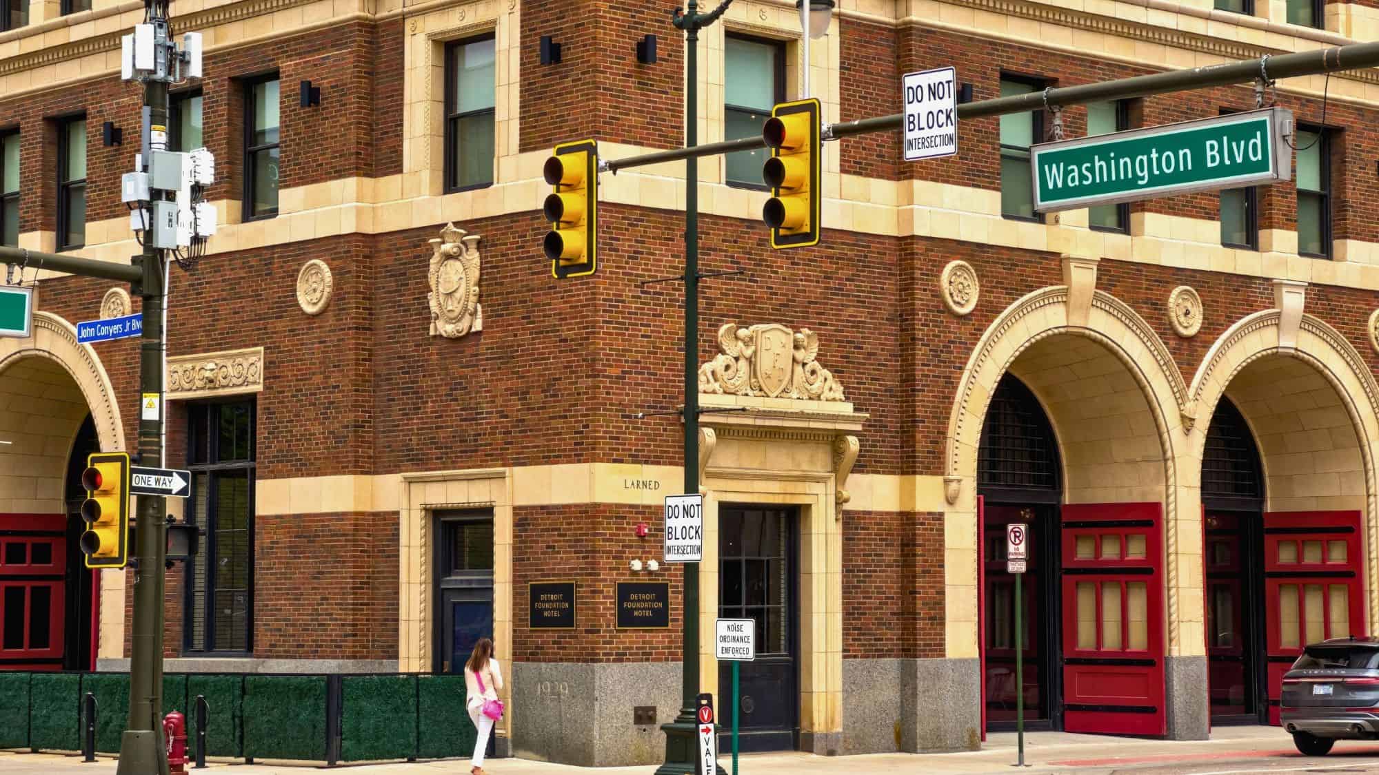 A historic brick building with ornate stone detailing on the corner of Larned and Washington Blvd in Detroit, with signage for the Detroit Foundation Hotel and red doors beneath grand arches.