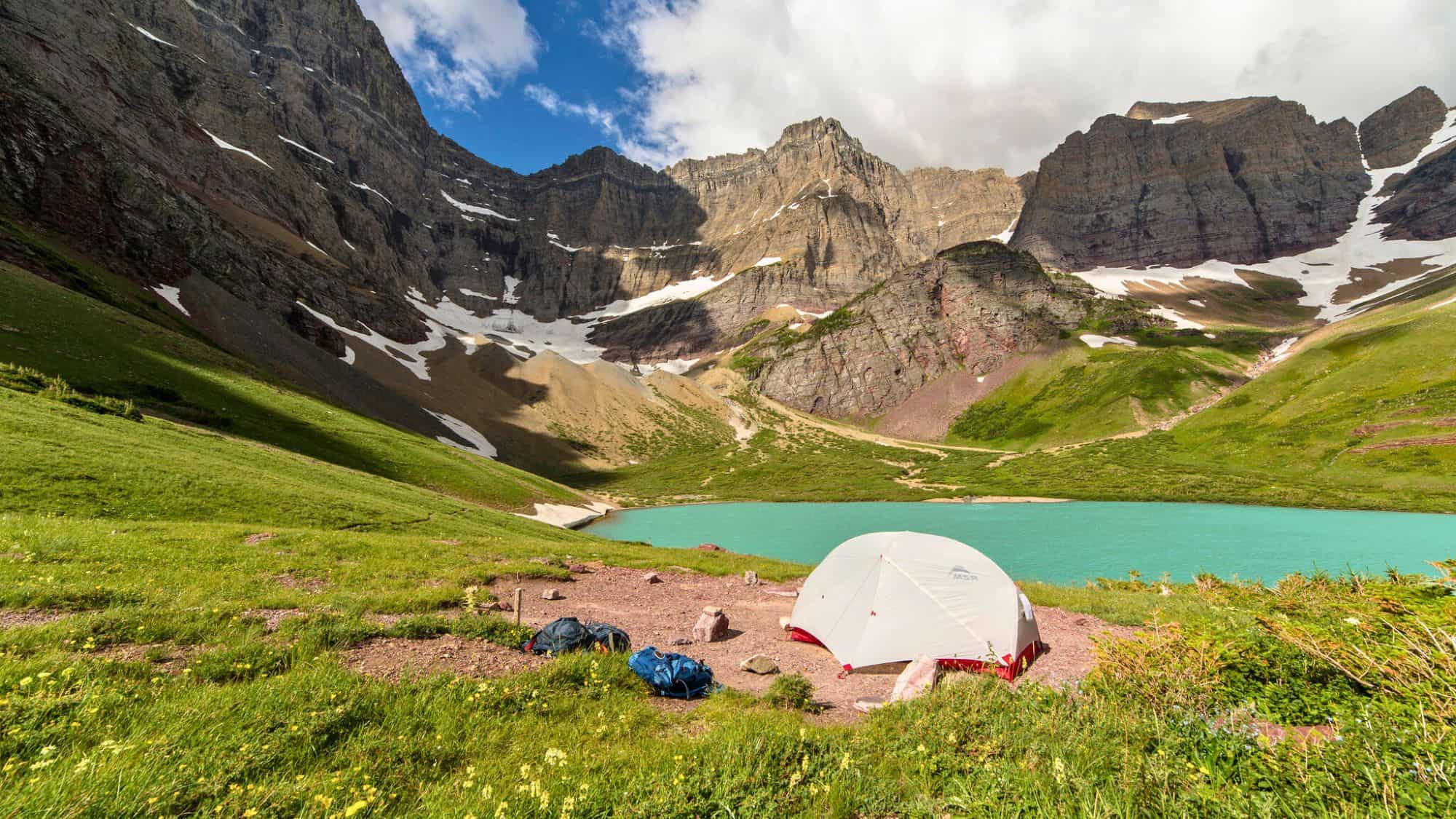 A small white tent is pitched beside a vibrant turquoise alpine lake beneath towering, rugged mountains dotted with patches of snow.