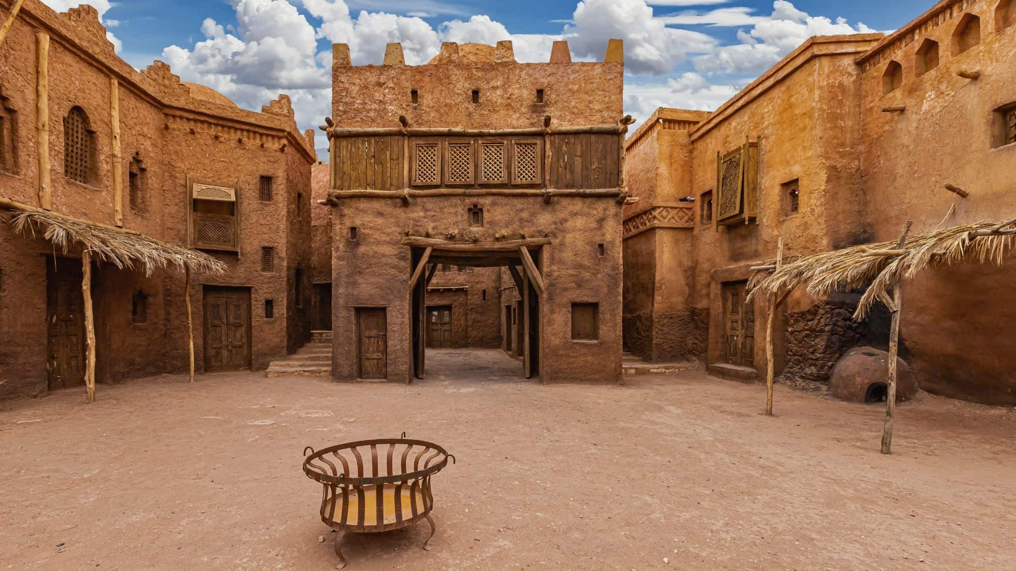 A traditional earthen Kasbah courtyard with wooden accents, rustic doors, and a central fire pit, set against a partly cloudy sky—this is a famous film set in Ouarzazate, Morocco.